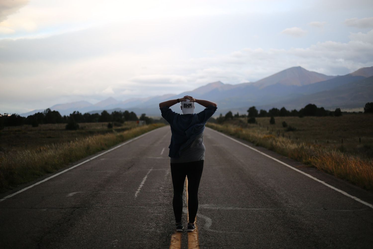 A silhouetted figure stands on an empty mountain road at dusk with distant peaks in the background.