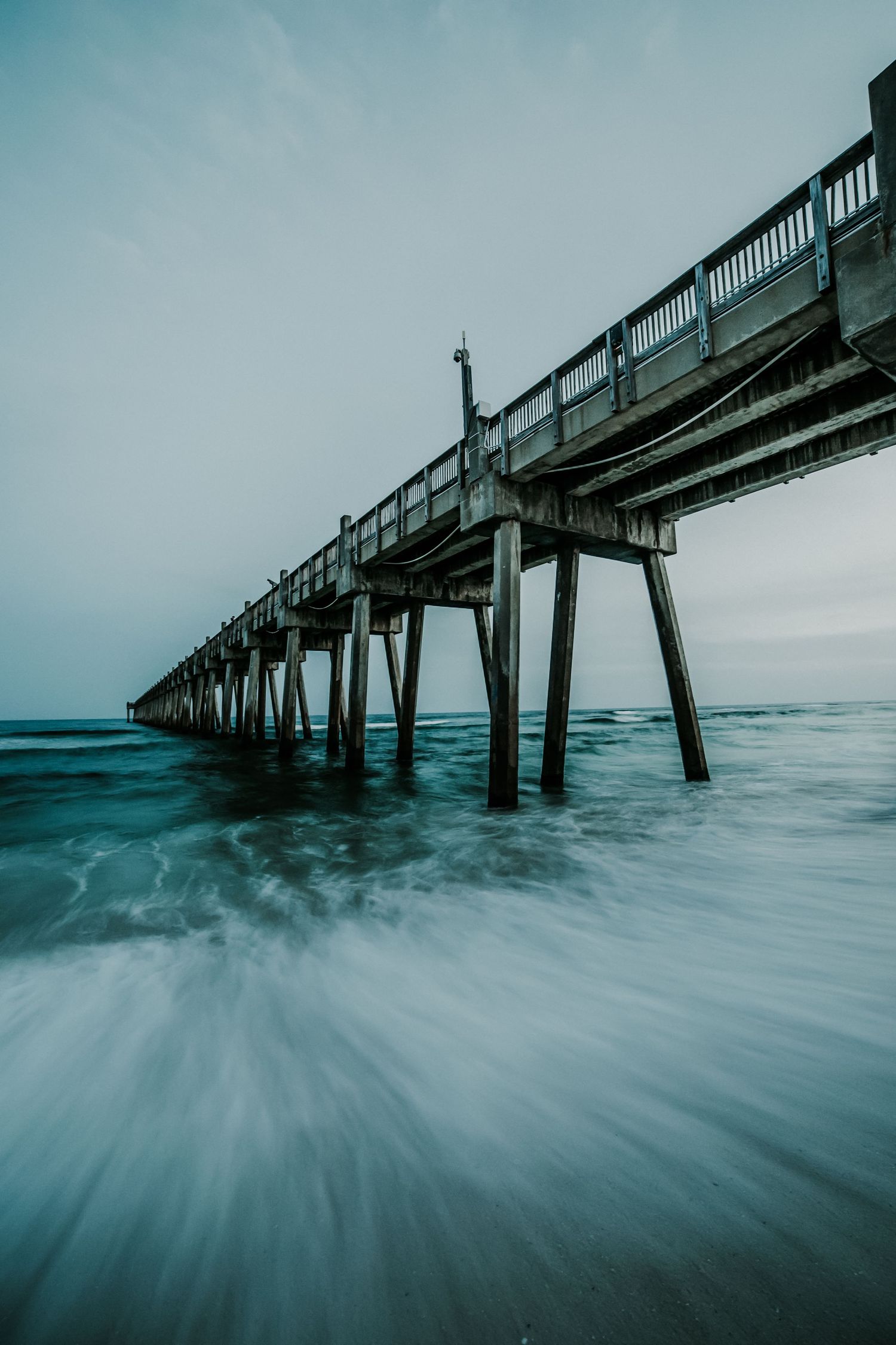 Long wooden pier stretches into turquoise ocean waters with dramatic long exposure waves creating a misty ethereal effect.