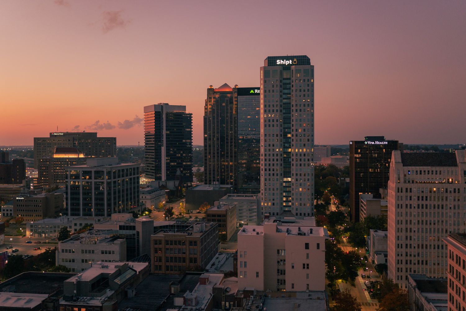 Panoramic cityscape at sunset showing modern buildings and skyscrapers against an orange and purple sky.