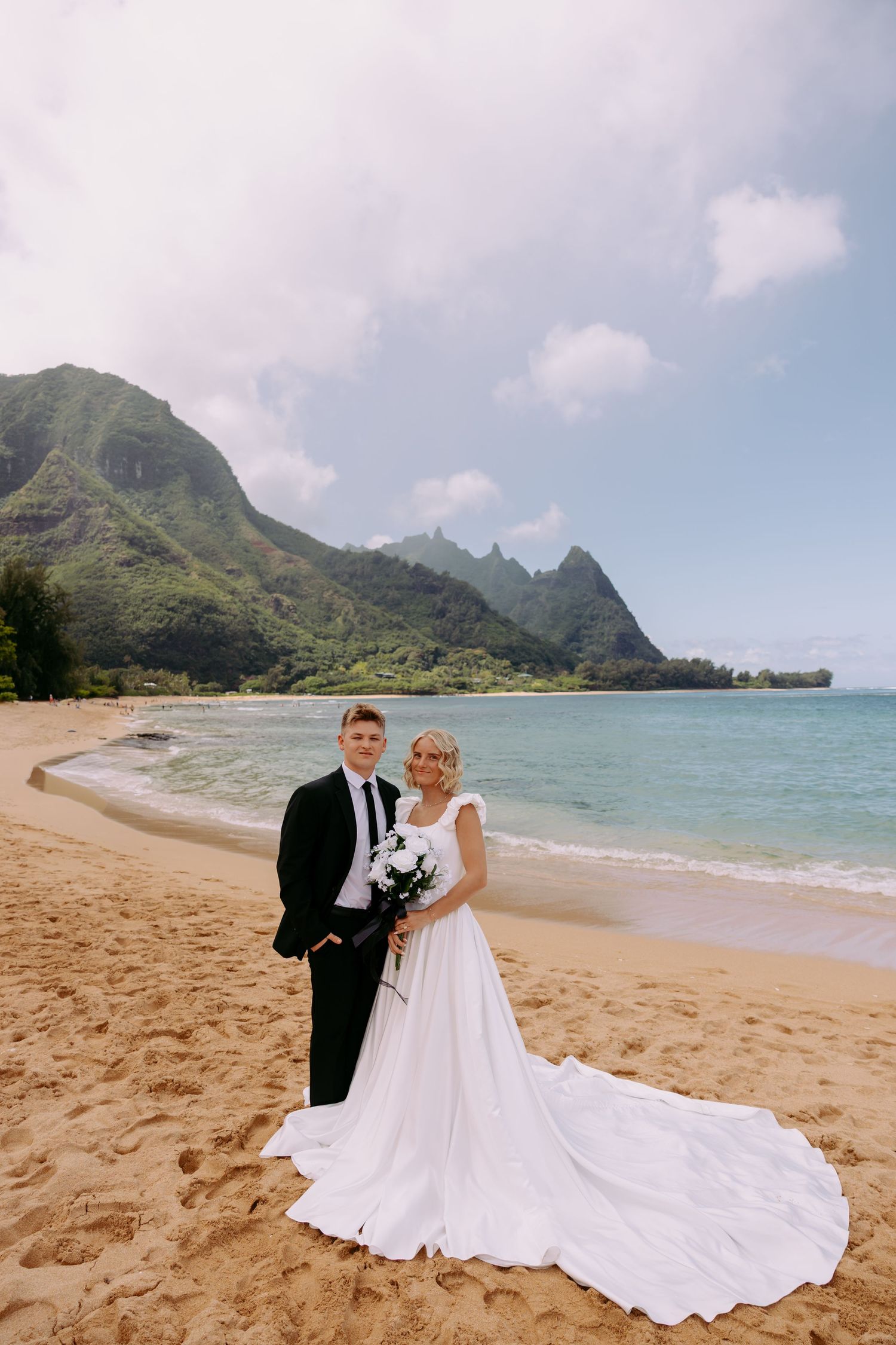 Bride's flowing white dress trails in sand as couple poses on scenic Hawaiian beach.