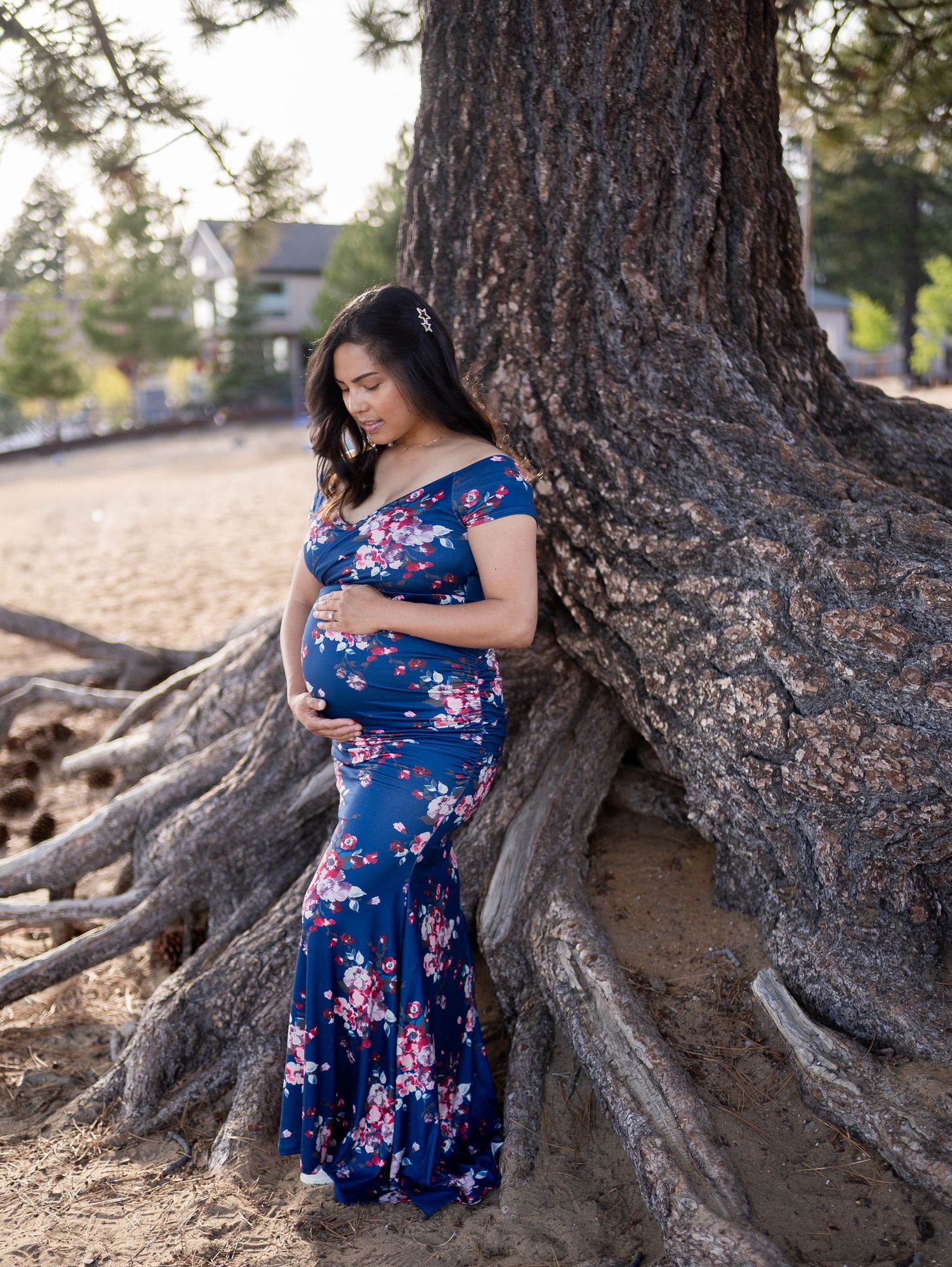 A person in a blue floral dress poses gracefully against the exposed roots of a large tree in a natural outdoor setting.