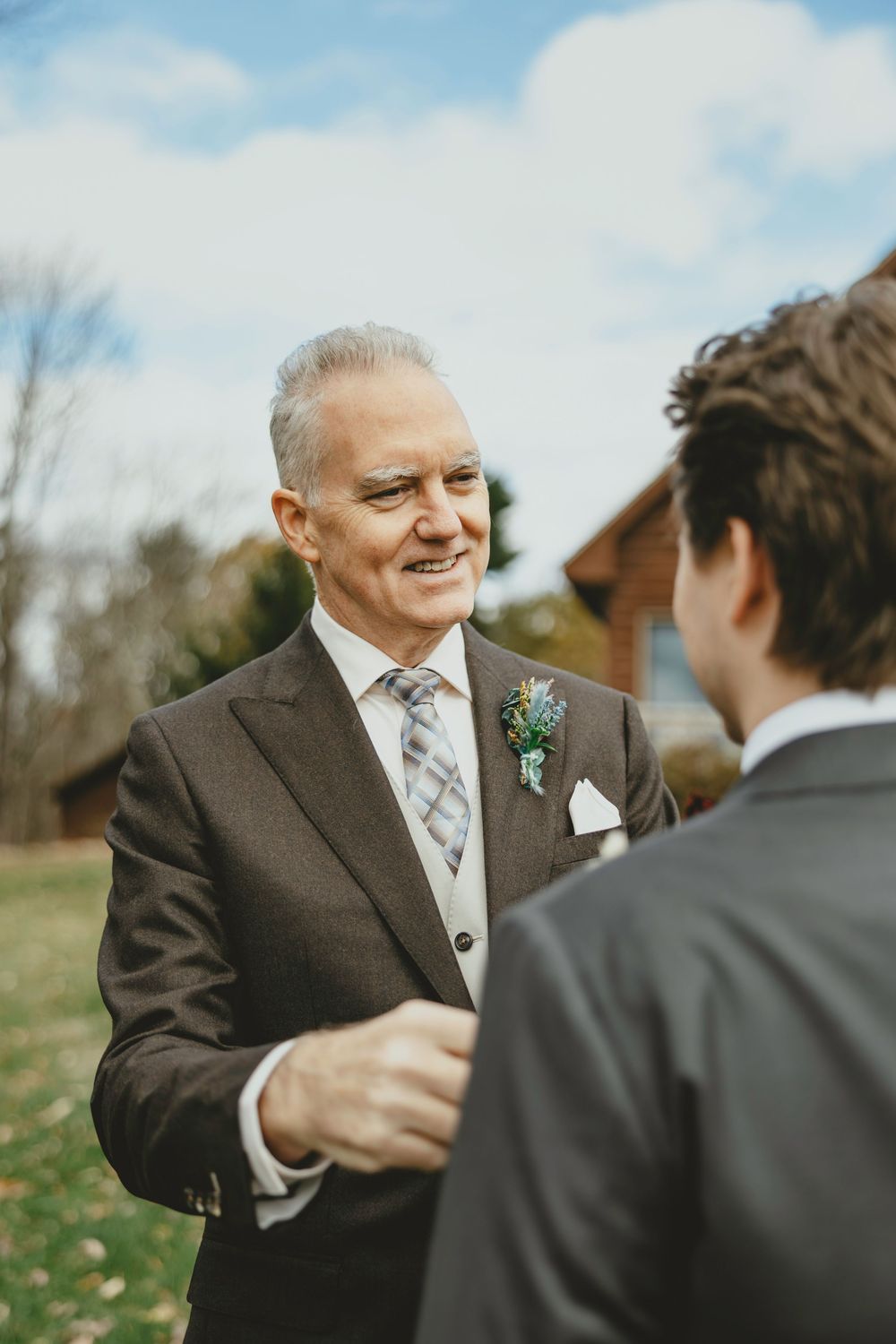 Two men in formal suits share an emotional moment outdoors during what appears to be a wedding celebration.