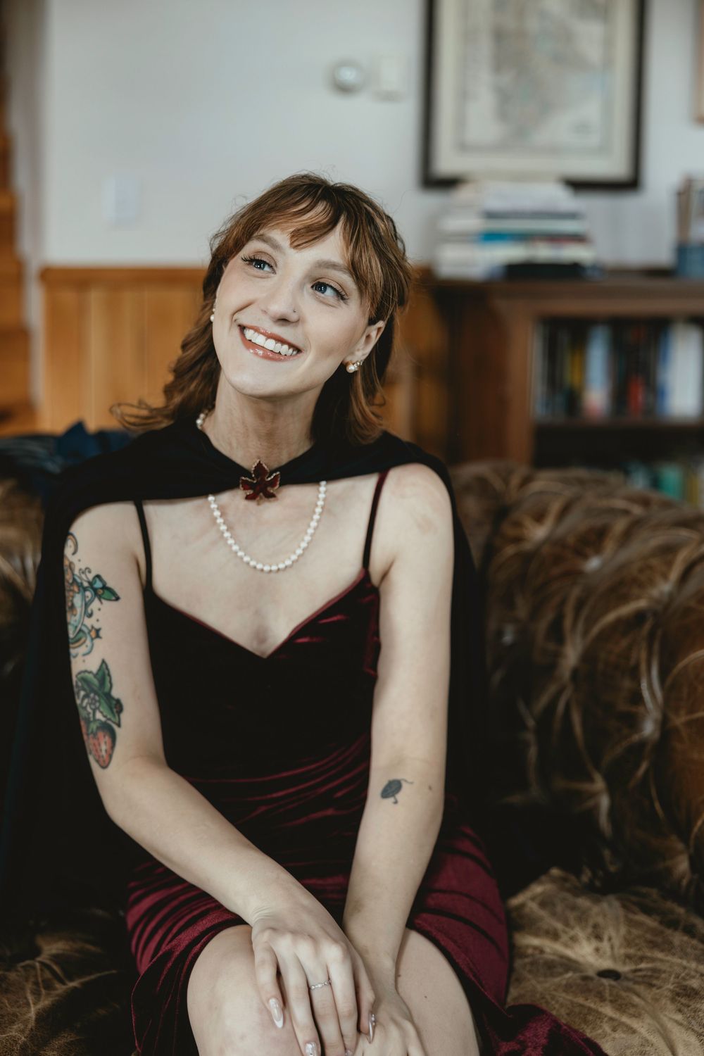 A woman in a black dress with tattoos sits on a couch wearing a pearl necklace and smiling warmly.