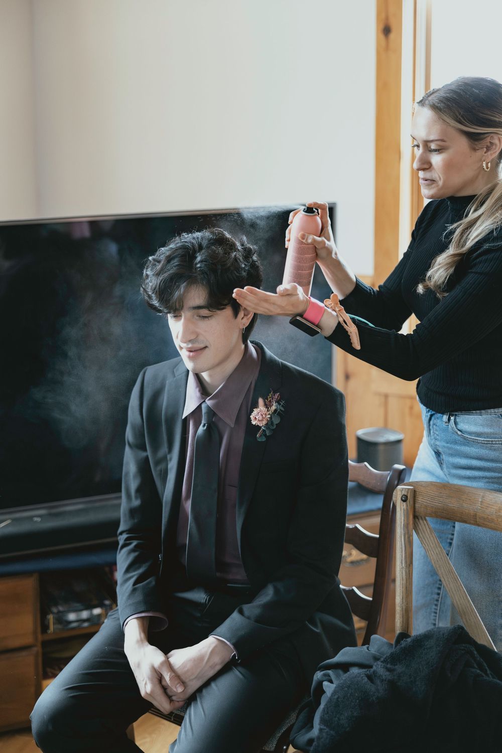 A person in a dark suit sits indoors while getting their hair styled for a formal event.