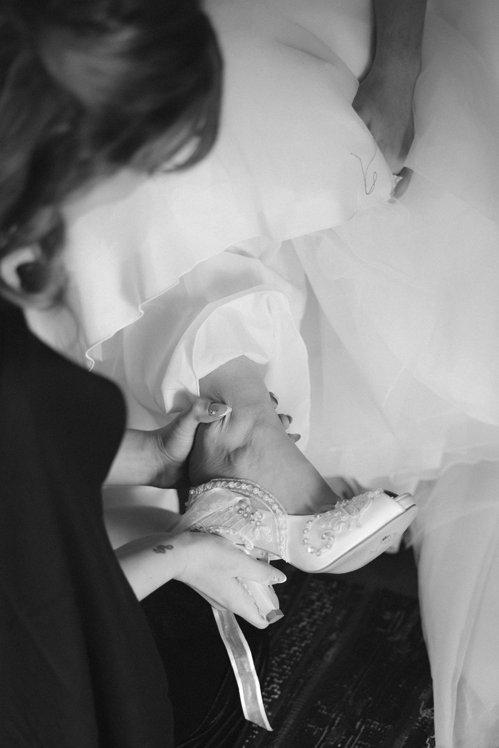 Black and white close-up of hands helping to put on an ornate bridal shoe with lace details.