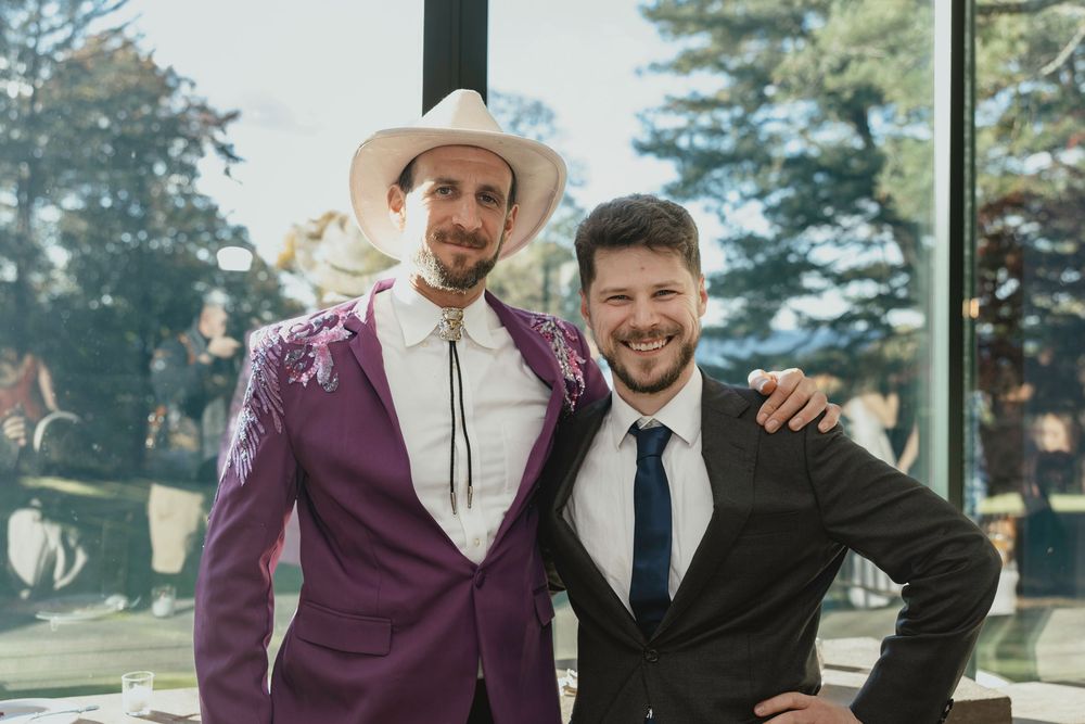 Wedding party members in purple and black formal attire pose together in front of large windows.