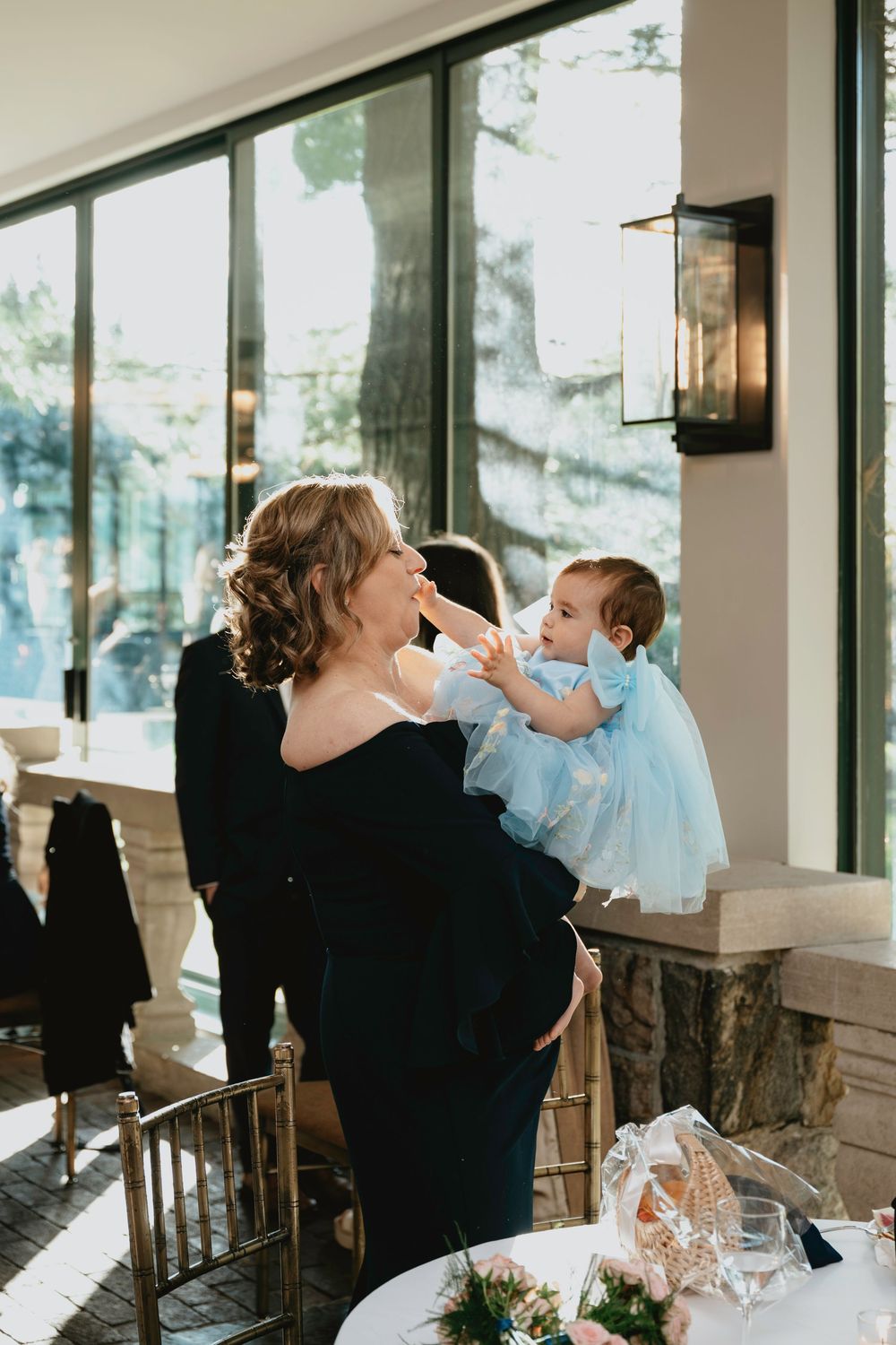 Wedding guest in black dress holds baby wearing light blue outfit in sunlit reception venue.