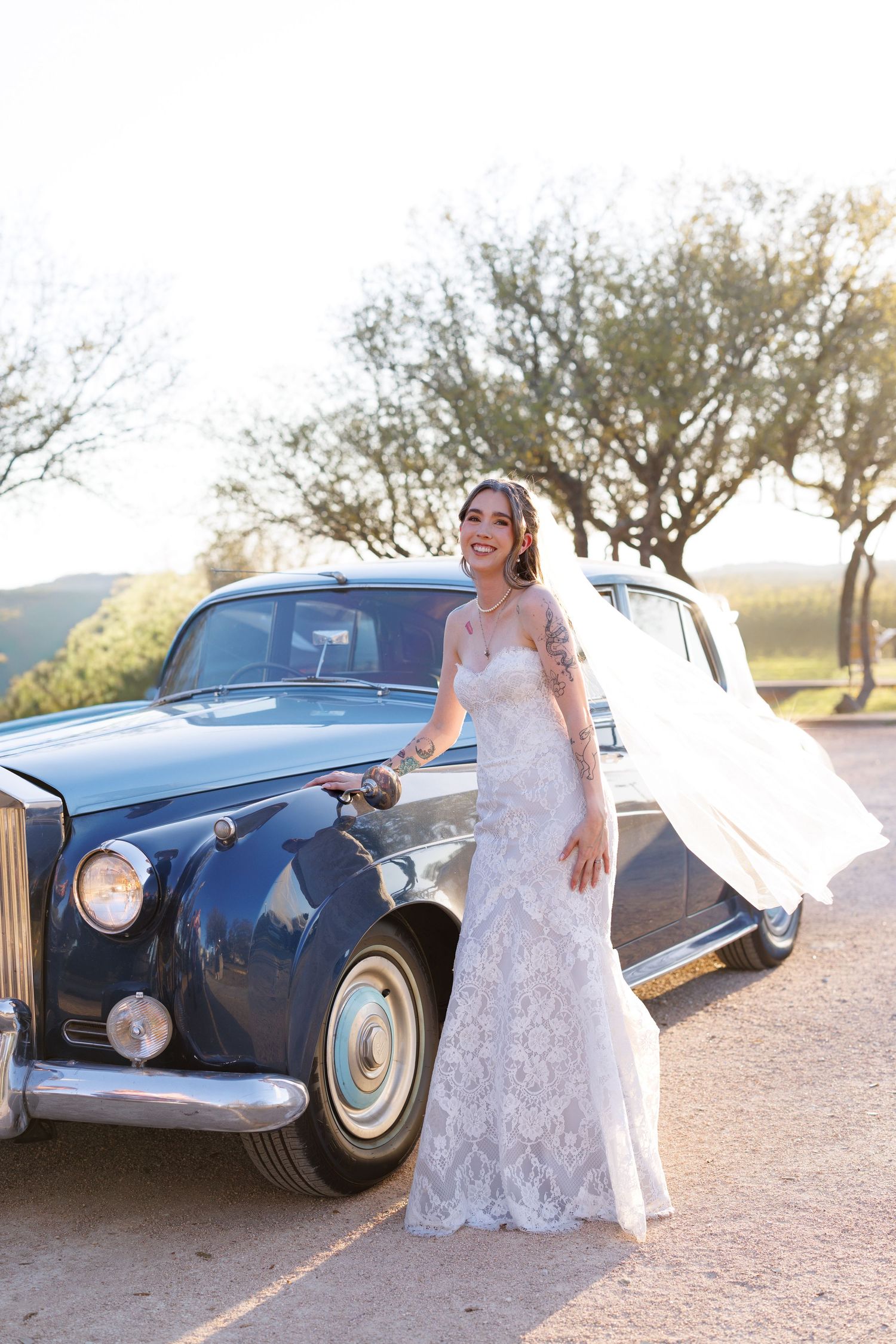 A white lace wedding dress photographed next to a classic blue vintage car during golden hour.