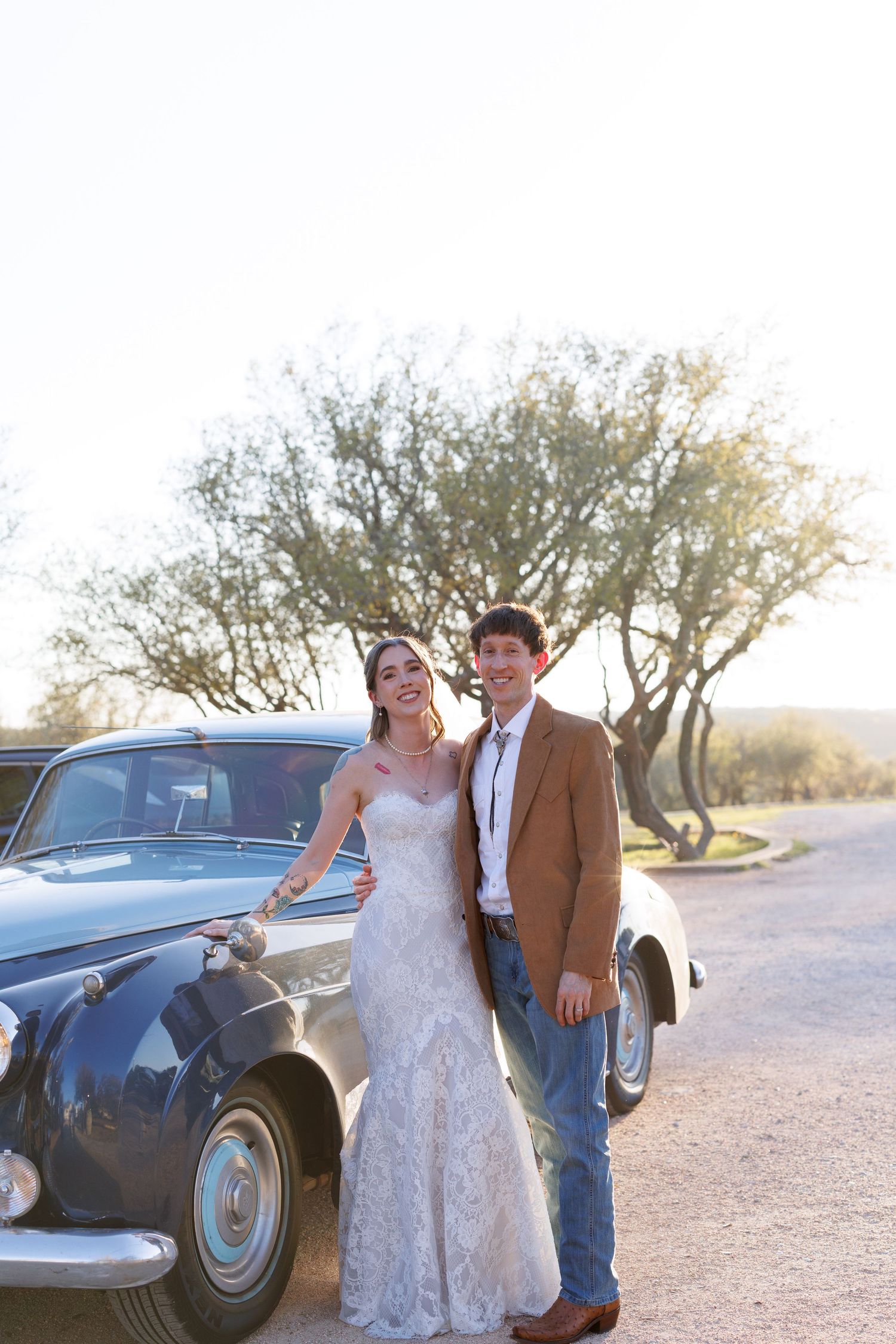 A couple poses next to a vintage car on a desert road with trees in the background during golden hour.