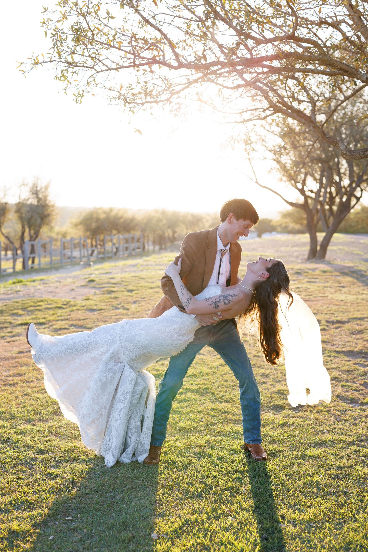 A romantic couple in wedding attire dances and embraces in a sunlit field with trees at golden hour.
