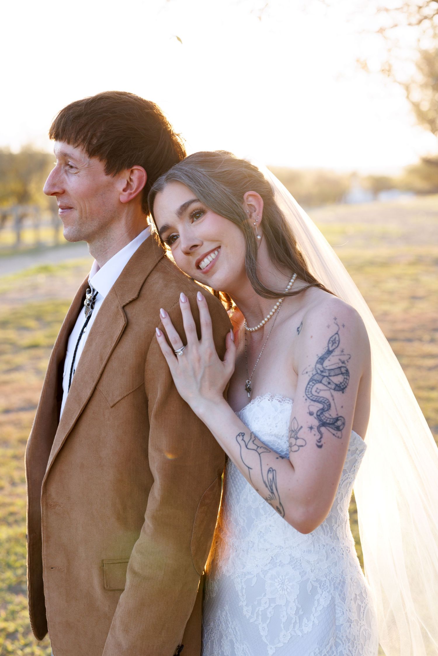 A romantic wedding couple embraces in golden sunlight, with the bride wearing a white dress and showing artistic tattoos.
