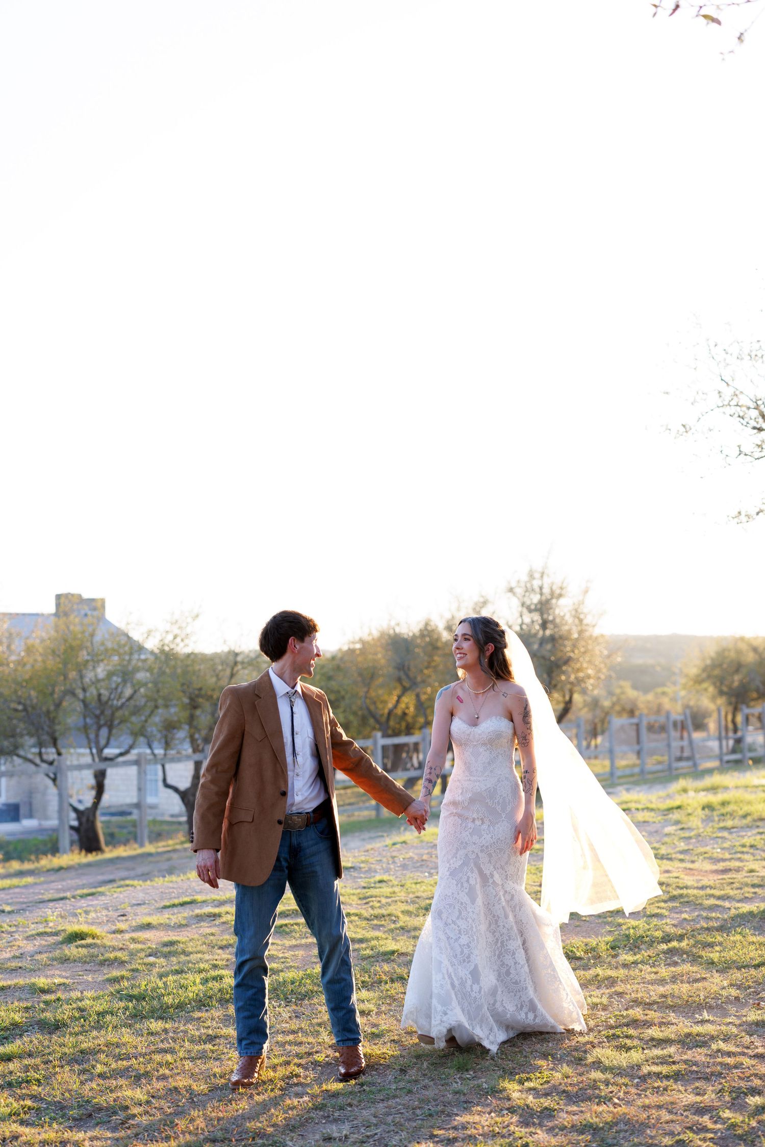 A couple in wedding attire walks together in a sunlit field during golden hour.