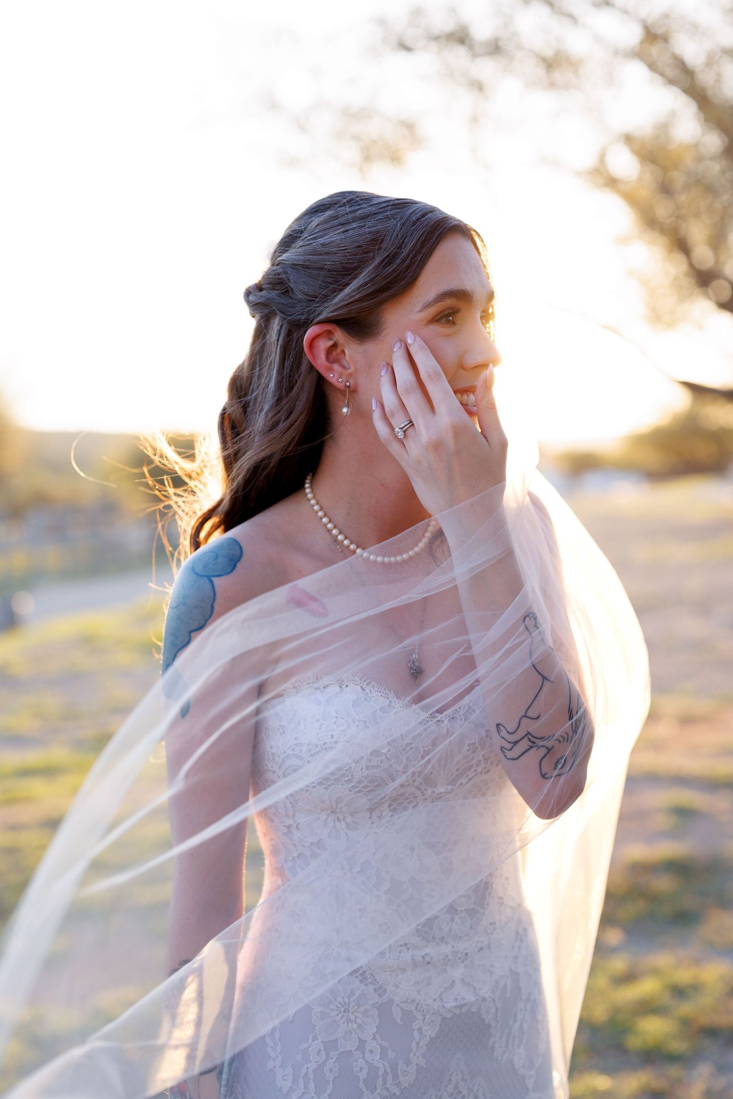 A bride in a white lace dress poses outdoors with a flowing veil during golden hour sunset.