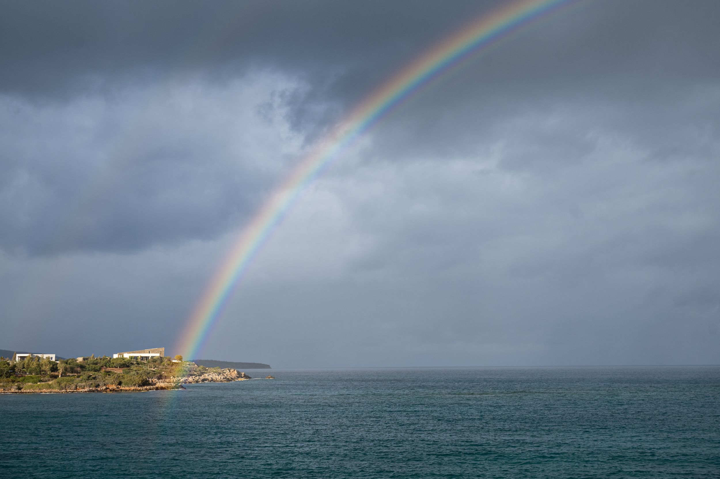 A vibrant double rainbow arches over a dramatic seascape with stormy clouds and coastal buildings in the distance.