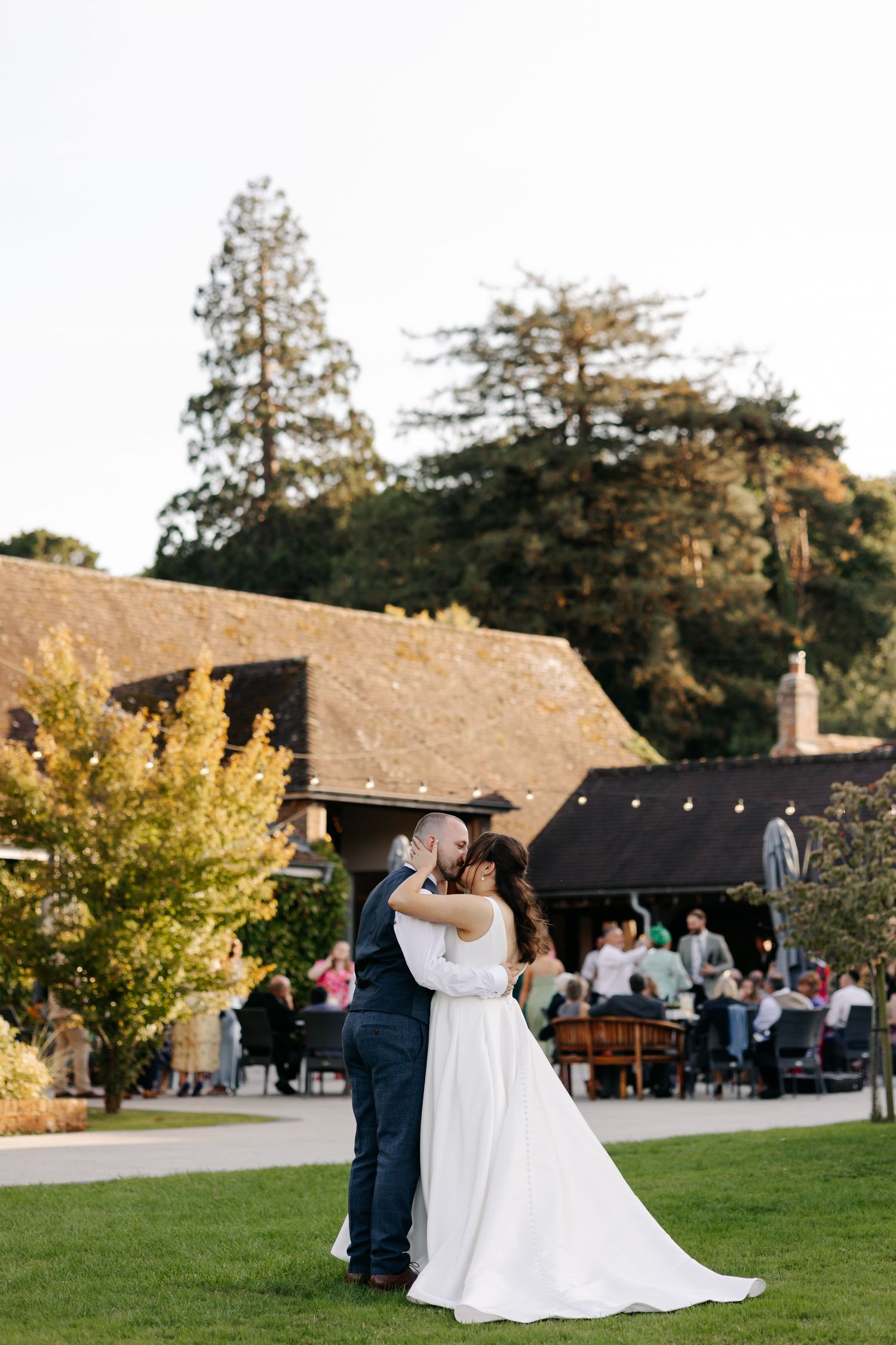 Romantic wedding couple shares their first dance outdoors on a grassy lawn at sunset with guests watching in background.