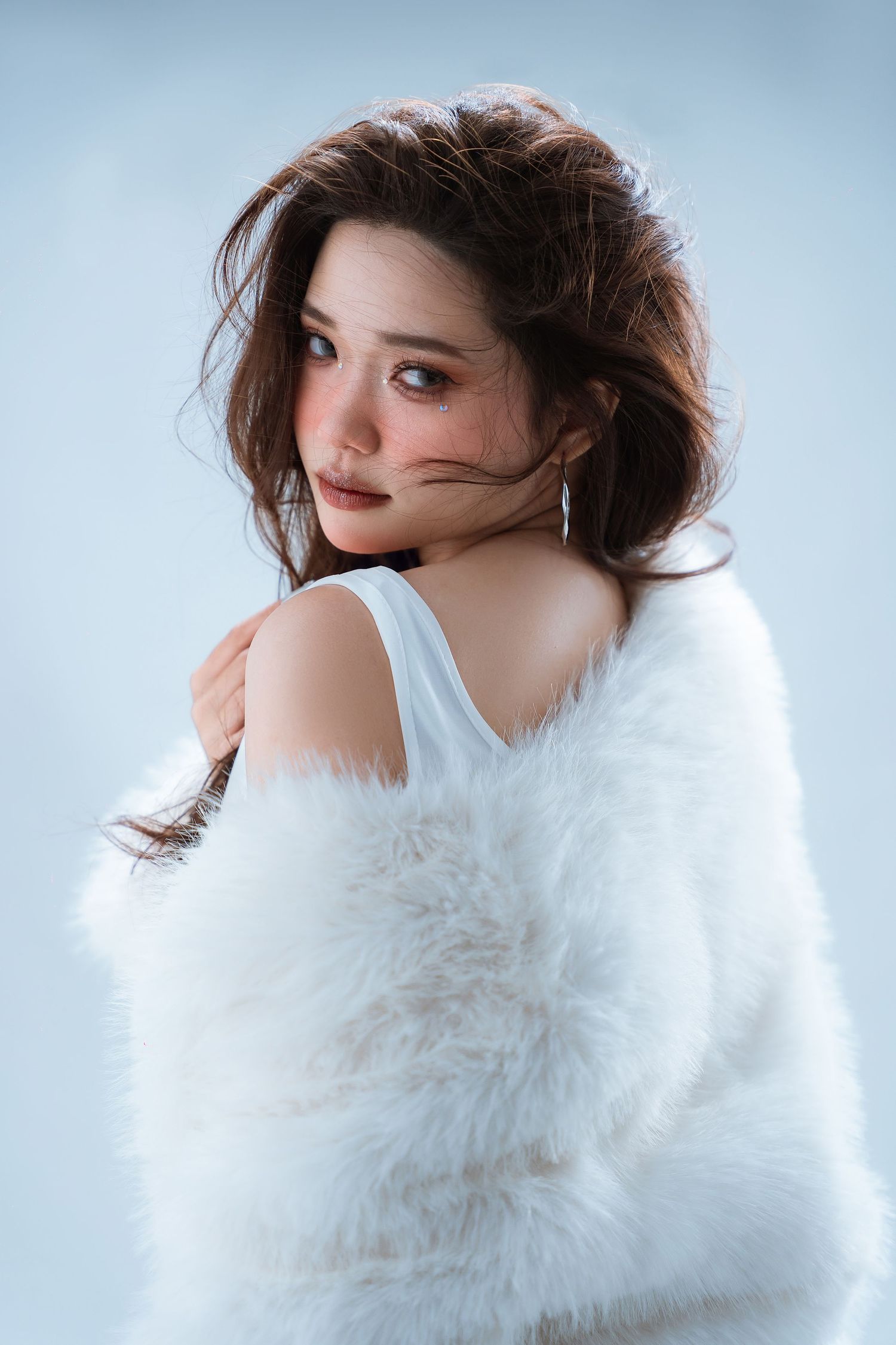 Glamorous portrait of someone in a white fur coat looking over their shoulder against a light blue backdrop.
