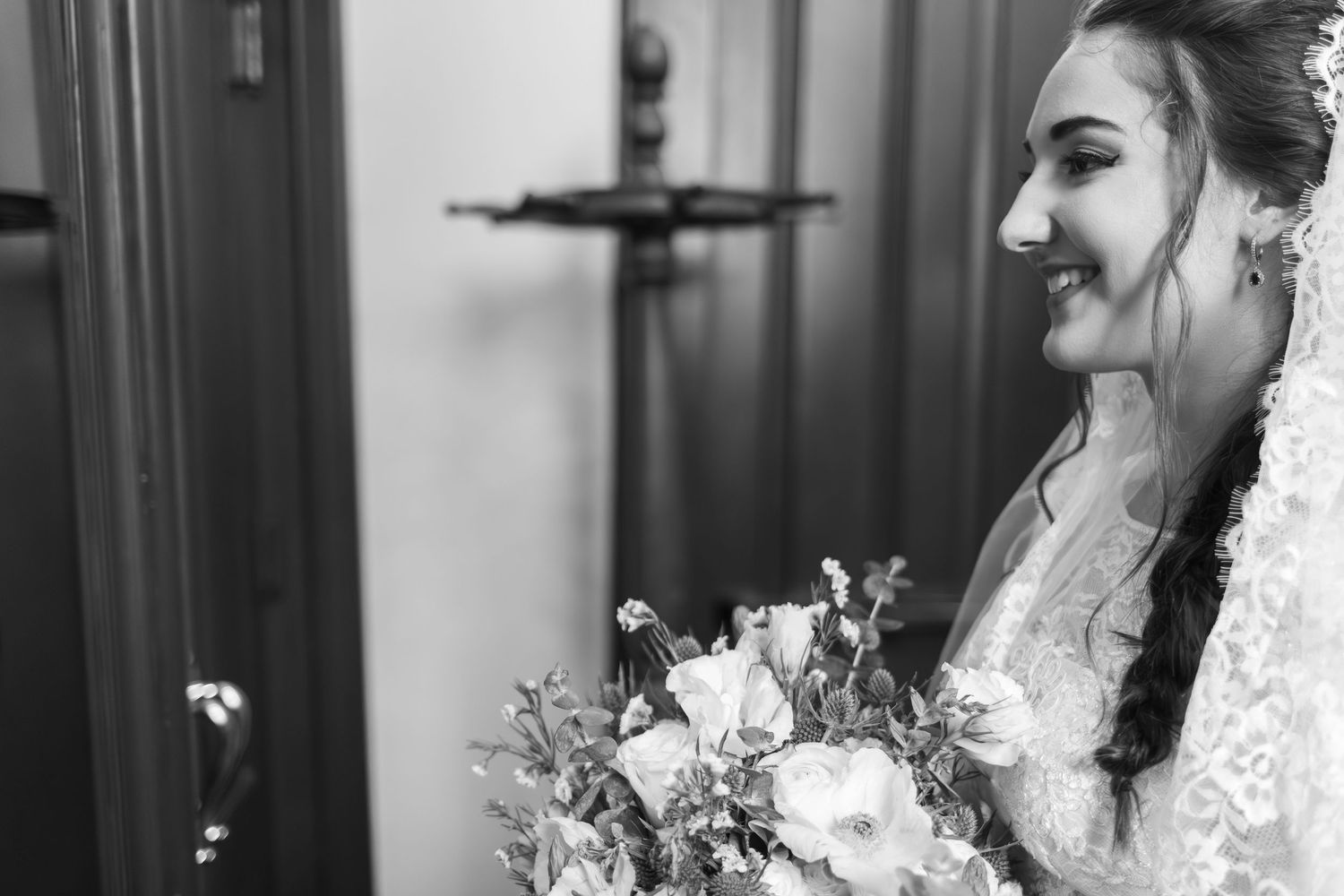 A bride in a lace wedding dress and veil laughs joyfully while holding a bouquet of white flowers.