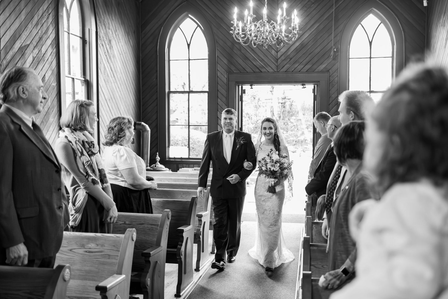A black and white photograph of a wedding ceremony inside a historic chapel with arched windows and a chandelier.