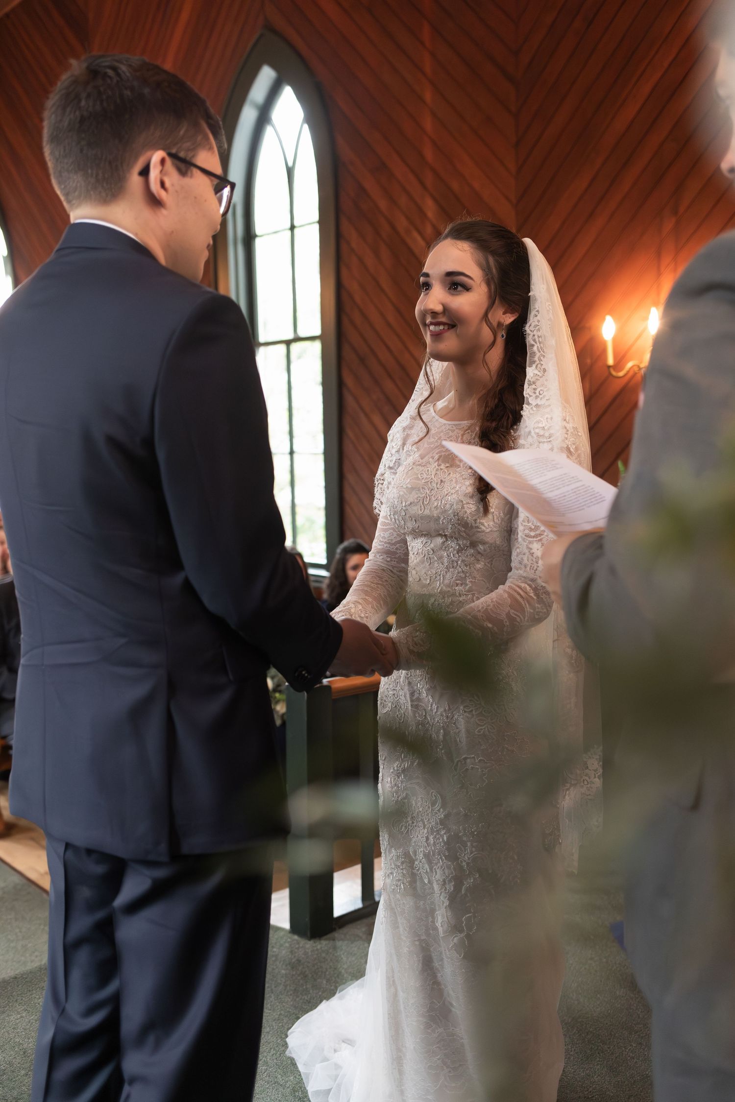 A bride and groom exchange vows during their wedding ceremony in a chapel with wooden walls and stained glass windows.