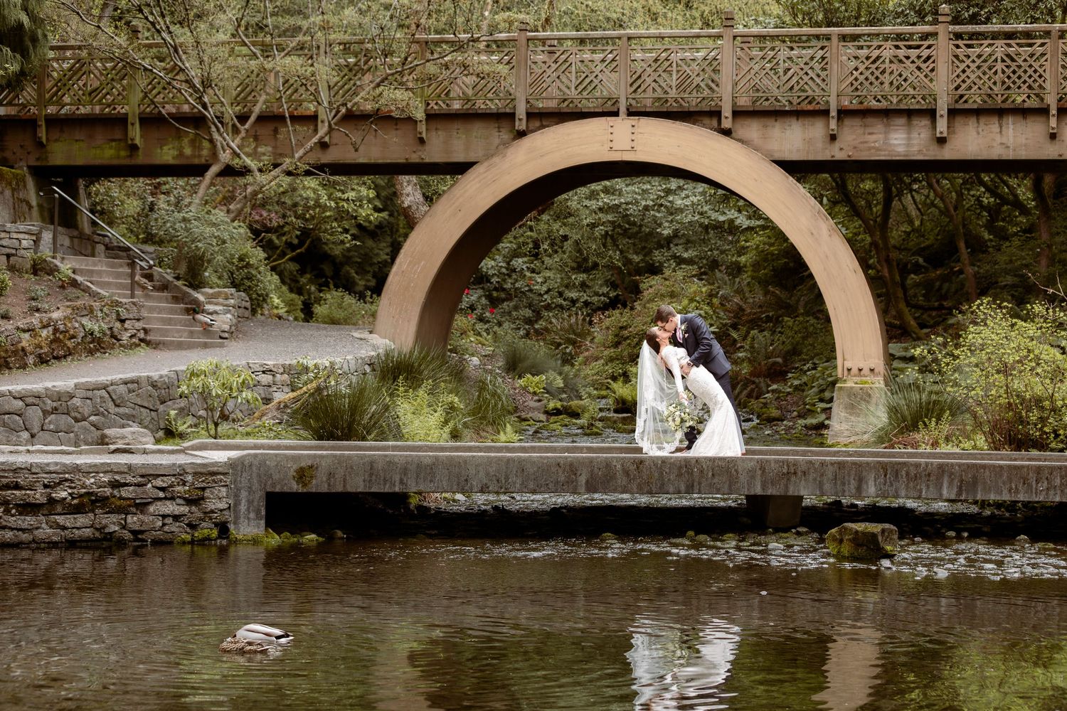 A couple in wedding attire poses on a stone bridge over a scenic stream in a lush garden setting.