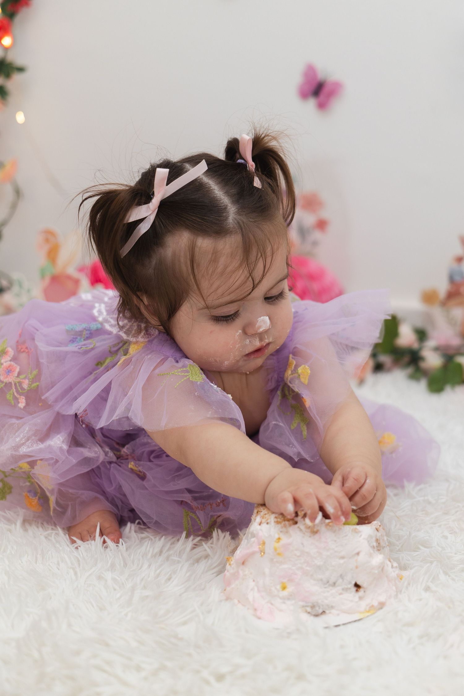 A small child in a purple tulle dress lies on a white fluffy rug surrounded by delicate floral decorations.