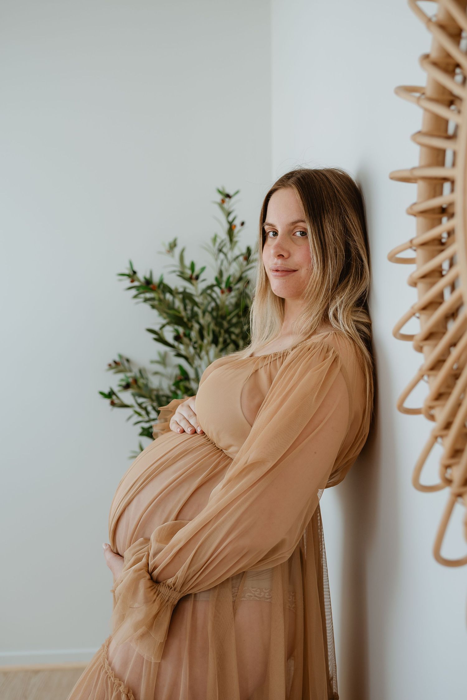 Pregnant woman in flowing beige dress stands against white wall with green plant in soft minimalist composition.