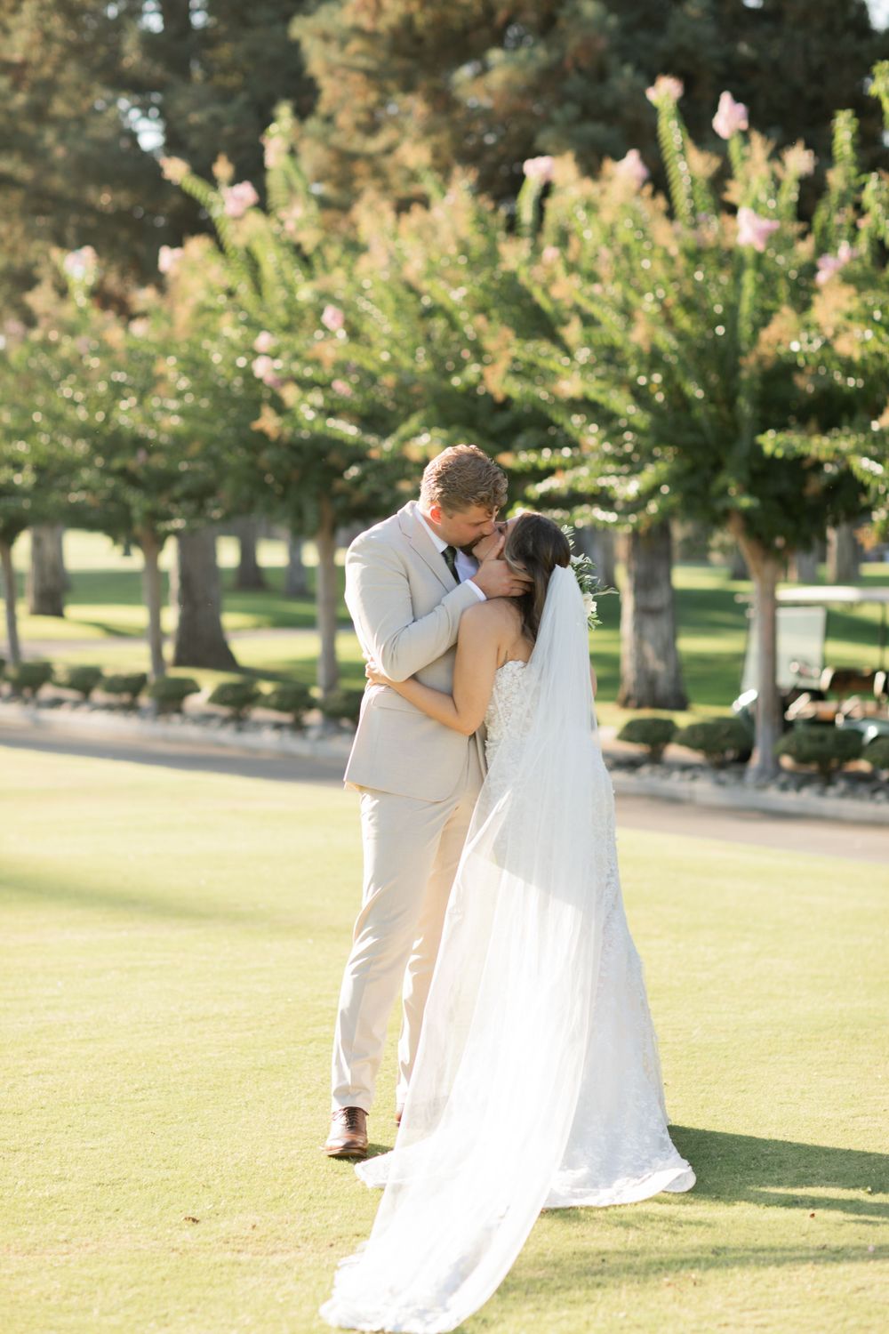 A romantic wedding couple shares a tender kiss on a sun-drenched golf course surrounded by trees.