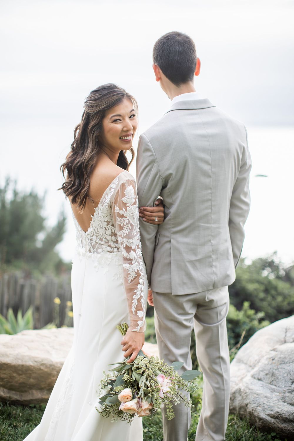A bride in a lace-sleeved wedding dress smiles while holding a bouquet next to her groom in a light gray suit outdoors.