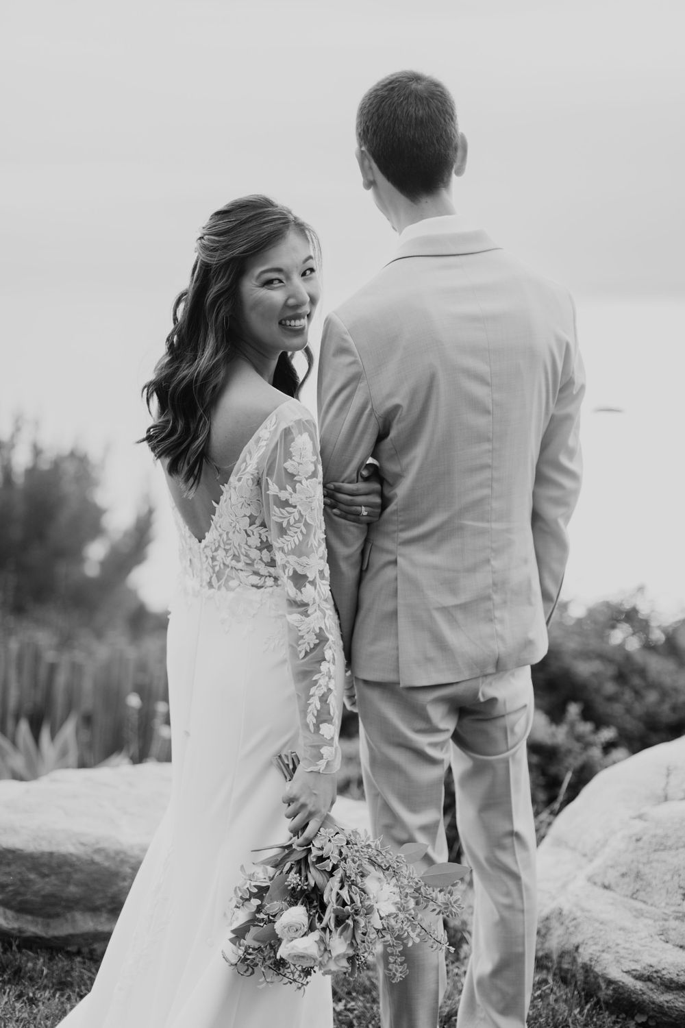 Black and white wedding photo of couple in lace gown and suit standing on rocks with bride looking over her shoulder.