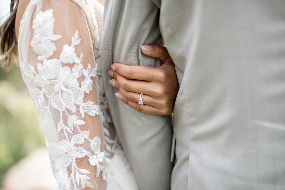 Close-up of hands with wedding rings against lace wedding dress sleeve and gray suit jacket.
