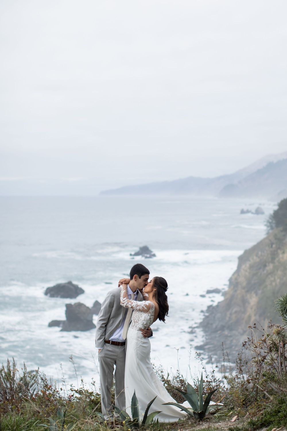 A couple embraces on a scenic coastal cliff overlooking the rugged Pacific Ocean shoreline with crashing waves and rocky outcrops.