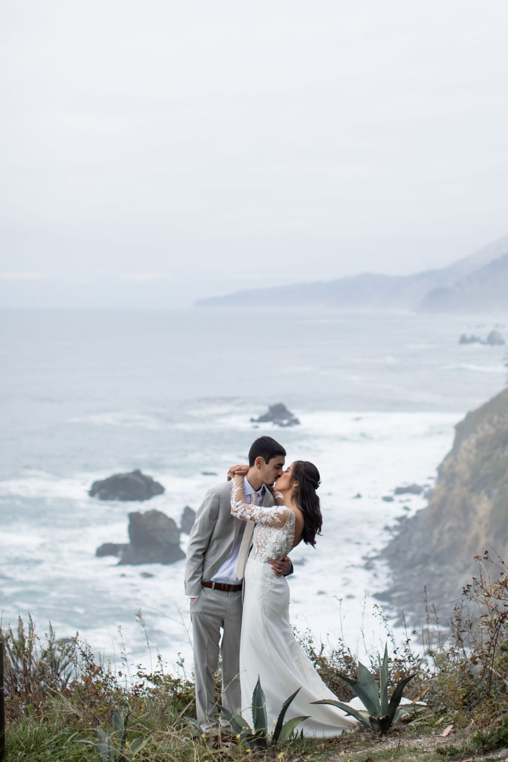 A couple in wedding attire embraces on a dramatic coastal cliff overlooking the misty Pacific Ocean and rocky shoreline.
