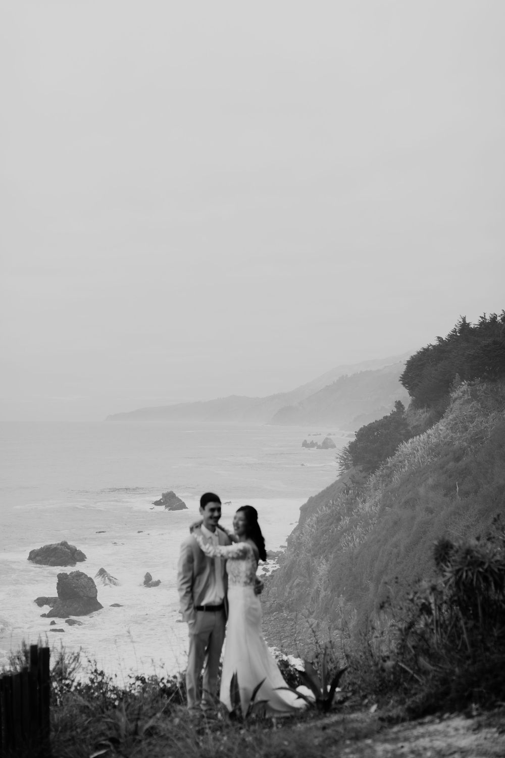 A romantic black and white photo series showing a couple embracing on a misty coastal cliff overlooking the ocean.