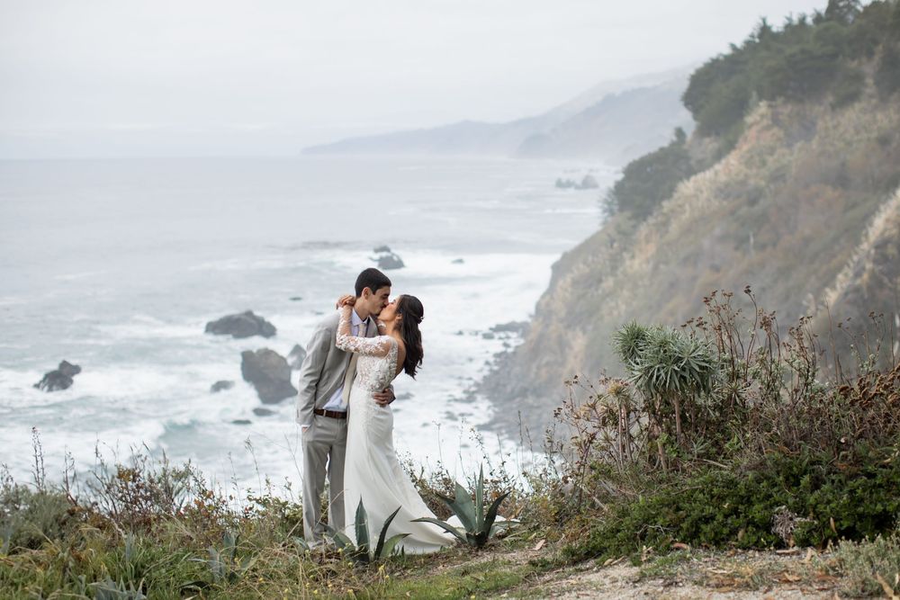 A couple embraces on a misty cliff overlooking the dramatic Big Sur coastline with crashing waves below.