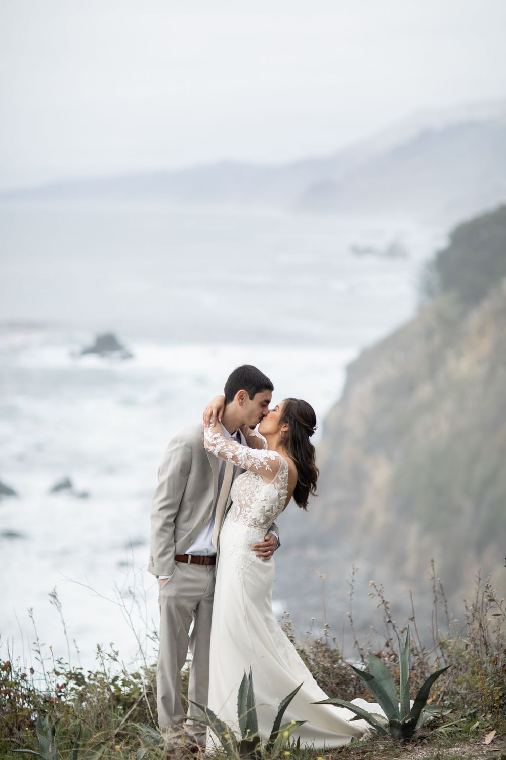 A couple shares a romantic moment on a misty coastal cliff overlooking the Pacific Ocean.