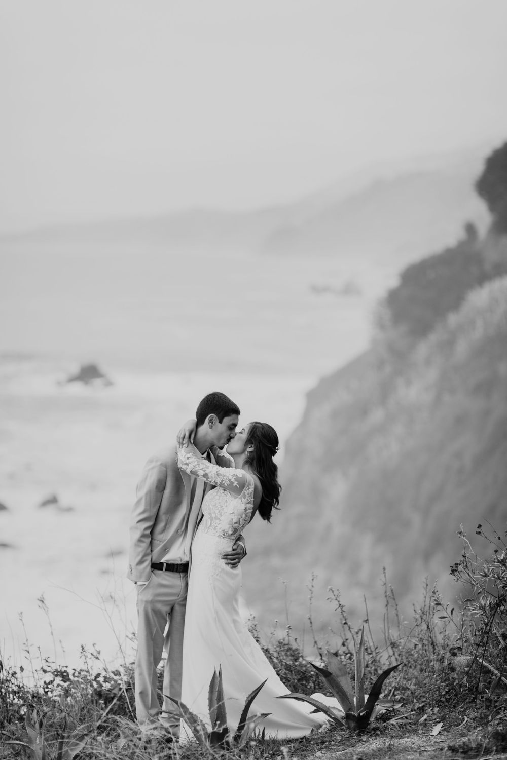 A romantic black and white photo series of a couple embracing on a coastal cliff with mountains and ocean in the background.