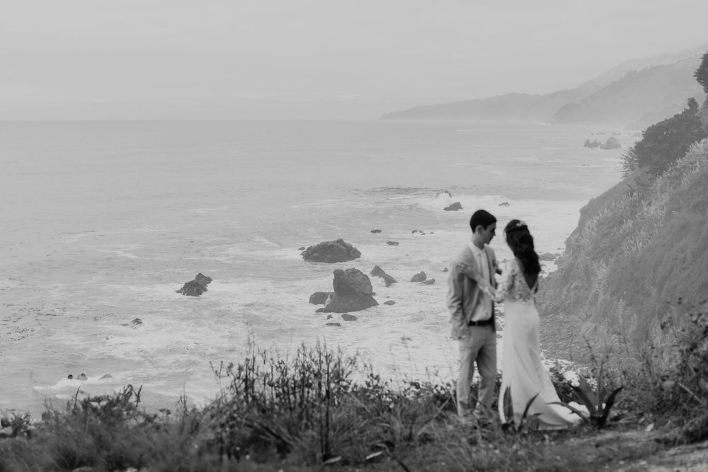 A couple stands on a misty coastal cliff overlooking rocky shores and ocean waves in black and white.