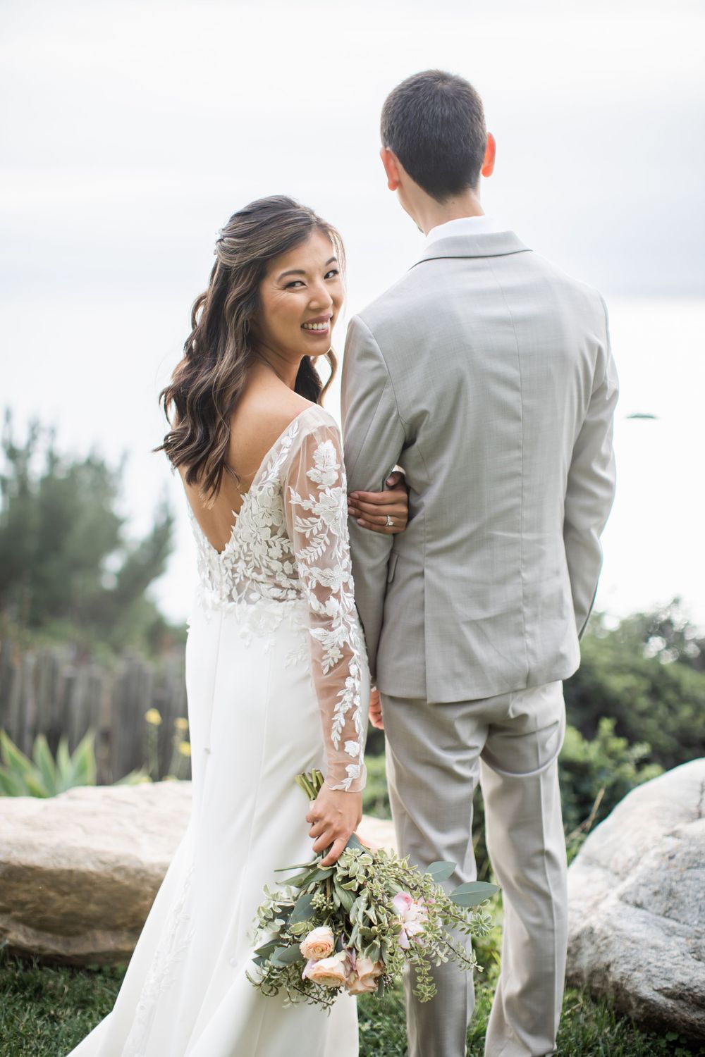 Bride in lace sleeve wedding dress looks back at groom in light gray suit while holding floral bouquet outdoors.