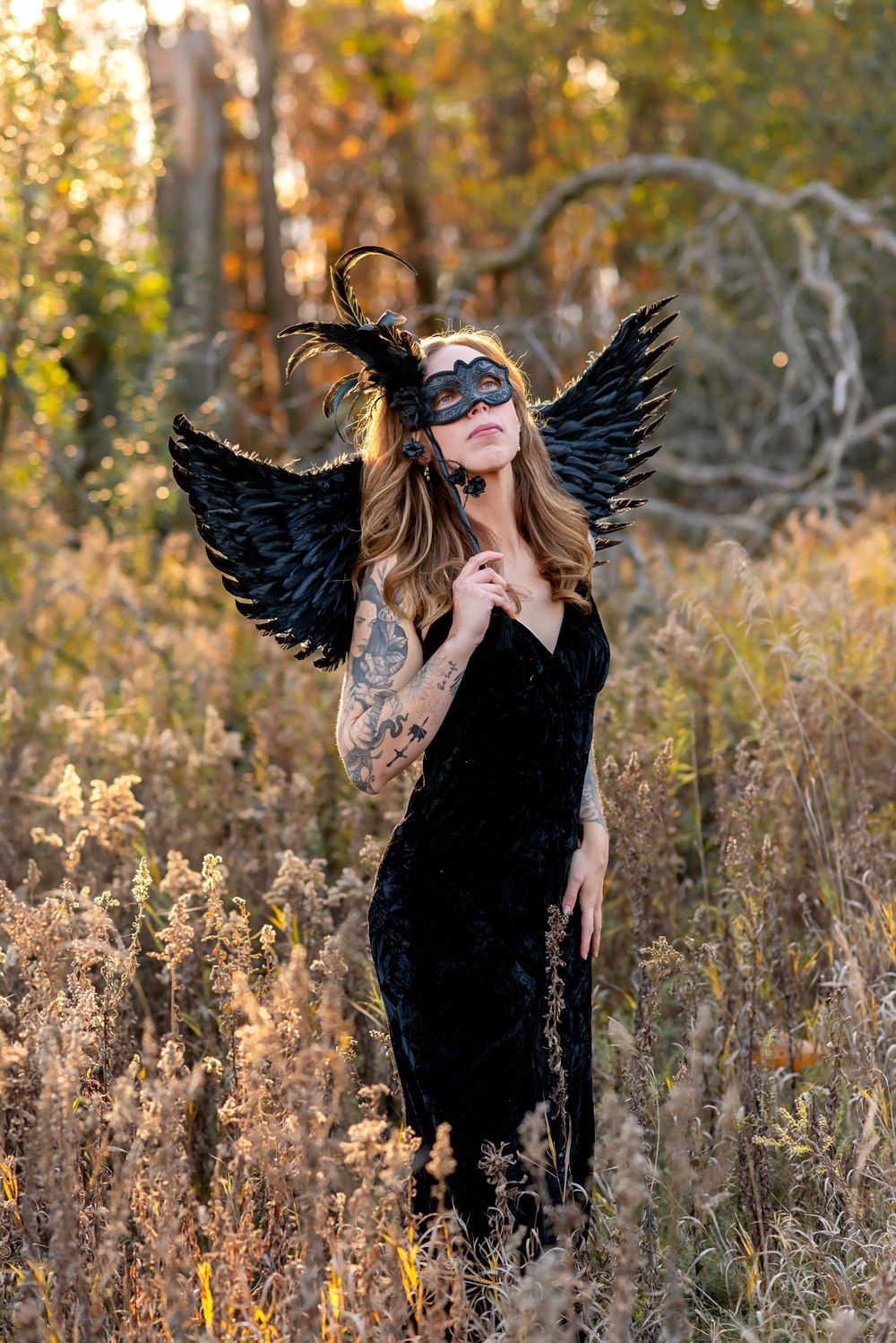 Woman in black velvet dress, feathered mask, and dark angel wings poses in golden autumn field at Komoka Provincial Park.