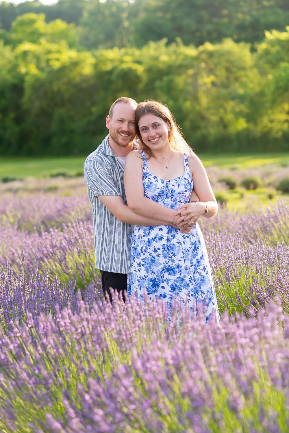 Couple embracing in a blooming lavender field, woman in blue floral dress smiling at camera during golden hour.