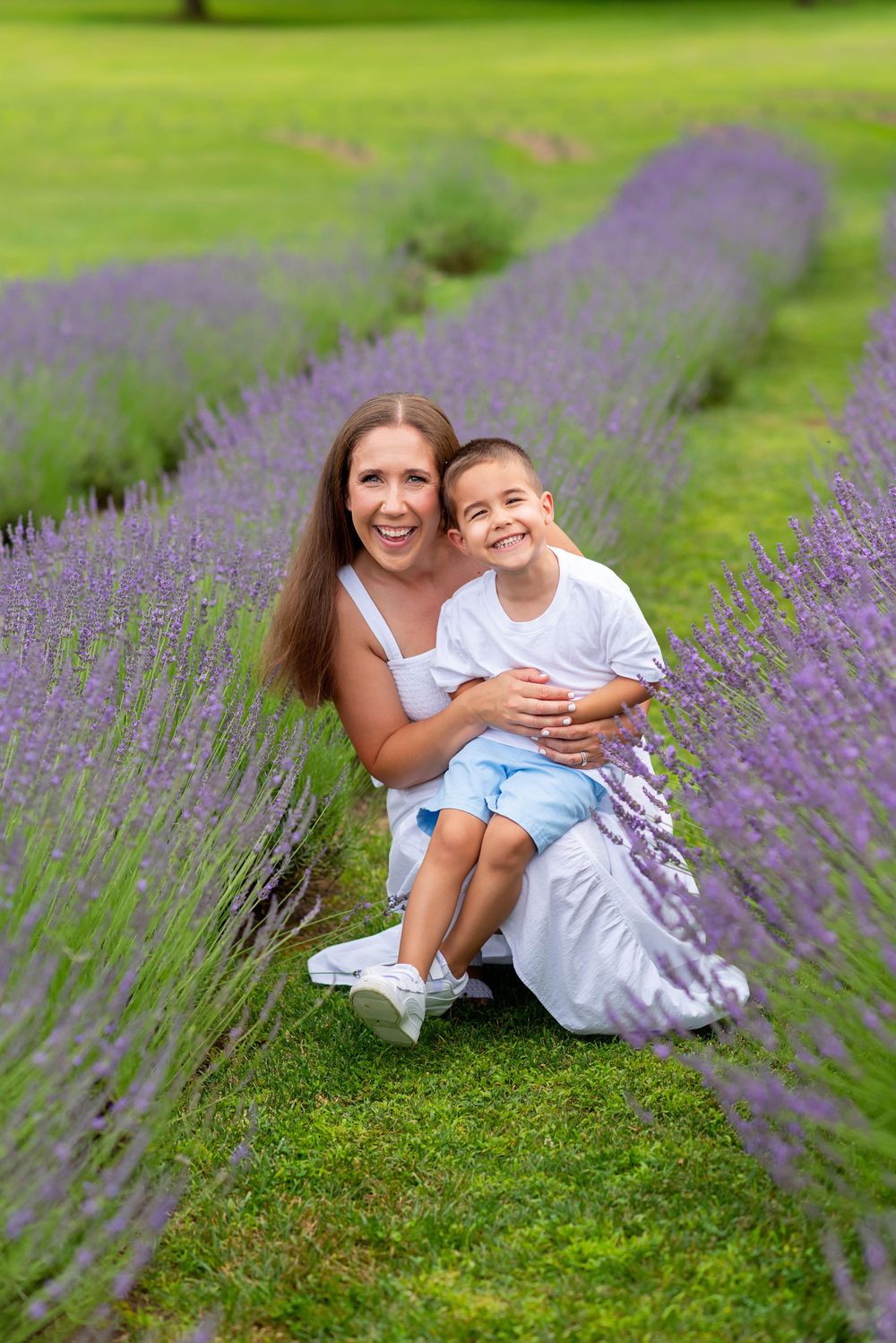 Happy mother hugging smiling boy in white shirt between rows of purple lavender flowers outdoors.