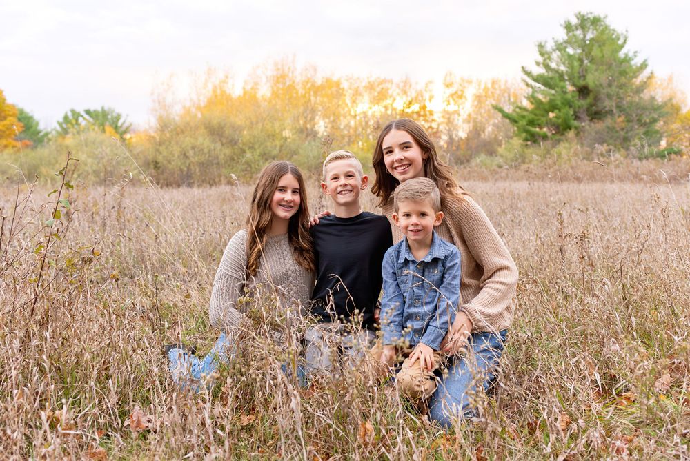 A smiling family of four poses together in a golden autumn field with colorful trees in the background at Komoka Provincial Park.
