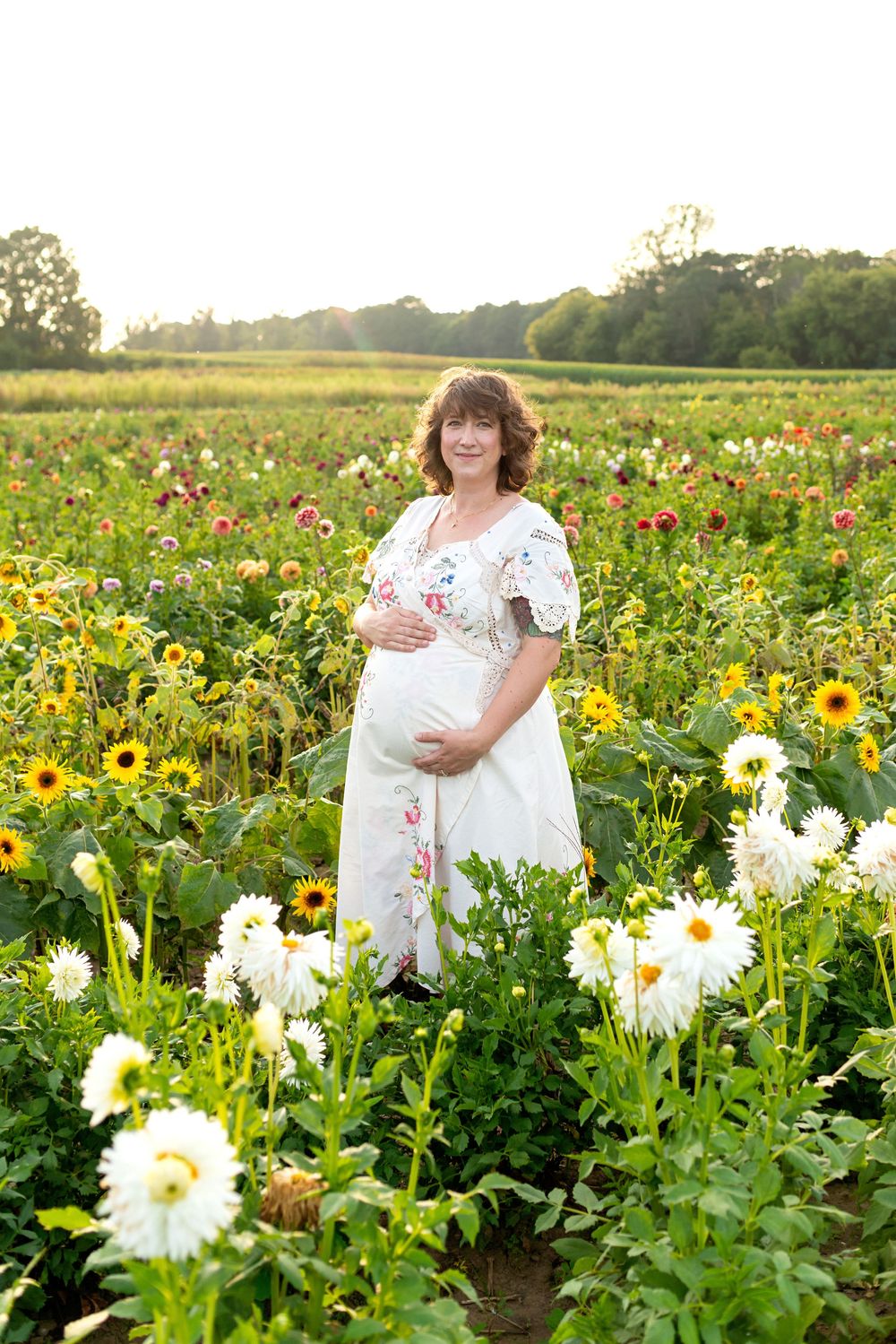 Pregnant woman in white floral dress standing in a vibrant flower field with sunflowers and dahlias at golden hour at the Flower lady and Ge