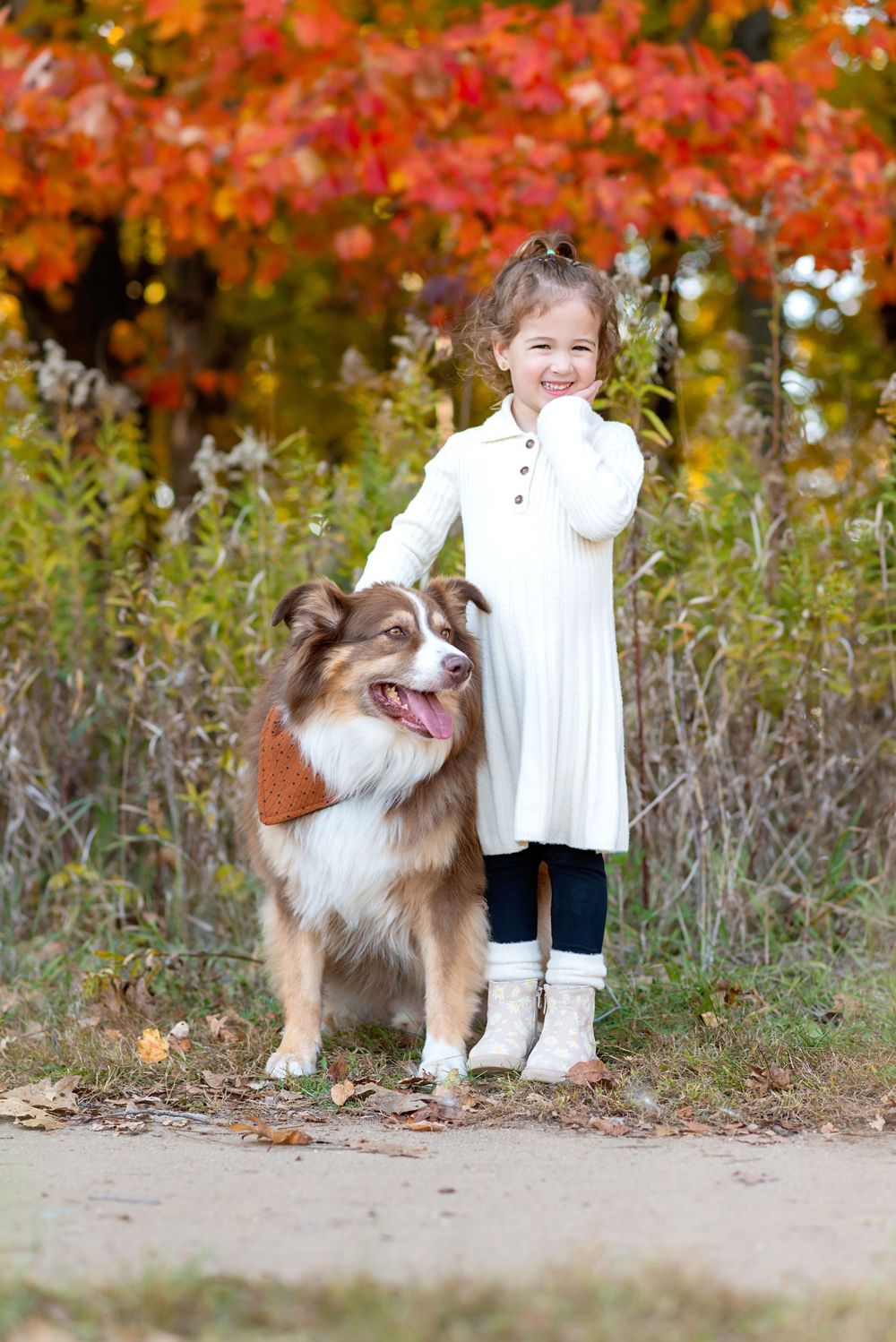 A smiling young girl in a white dress stands with her Australian Shepherd dog on a path surrounded by colorful autumn foliage at Komoka park