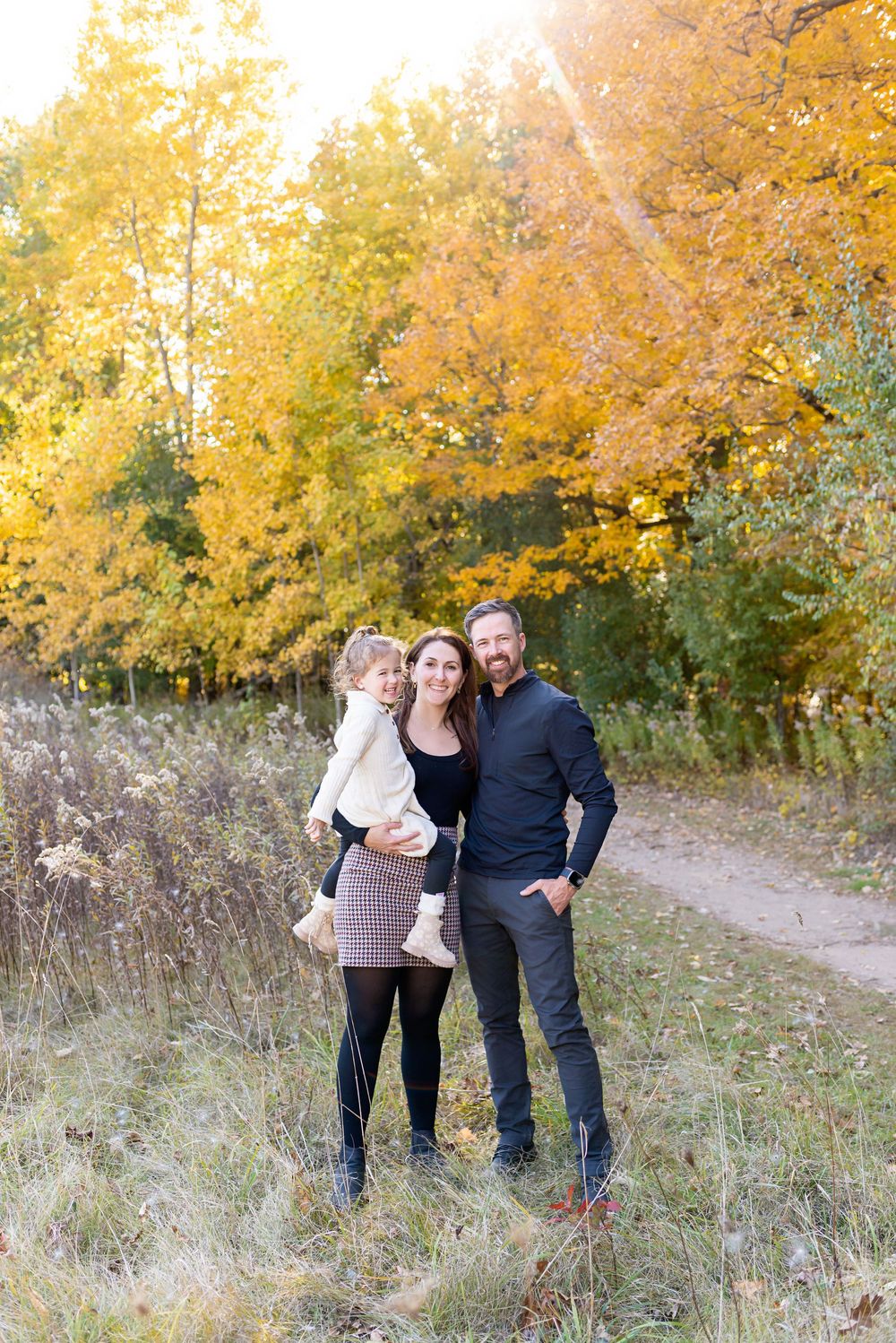Family of three posing outdoors among vibrant yellow autumn trees on a scenic dirt path in fall at Komoka Provincial Park.