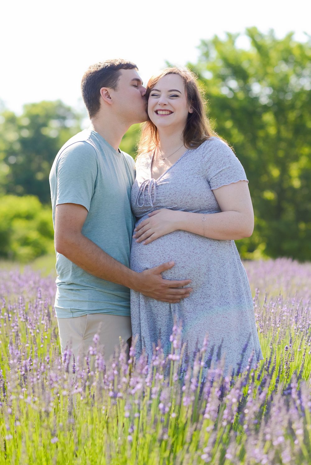 A man kisses a smiling pregnant woman in a blooming lavender field on a sunny day.