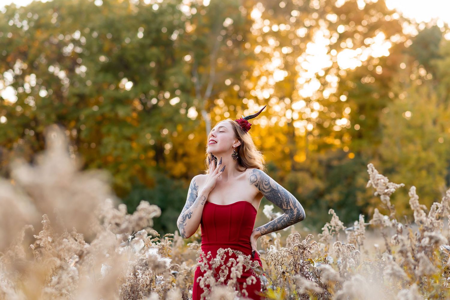 Woman in red dress with tattoos poses in golden field at sunset, wearing devil horns, surrounded by autumn trees at Komoka Provincial Park.
