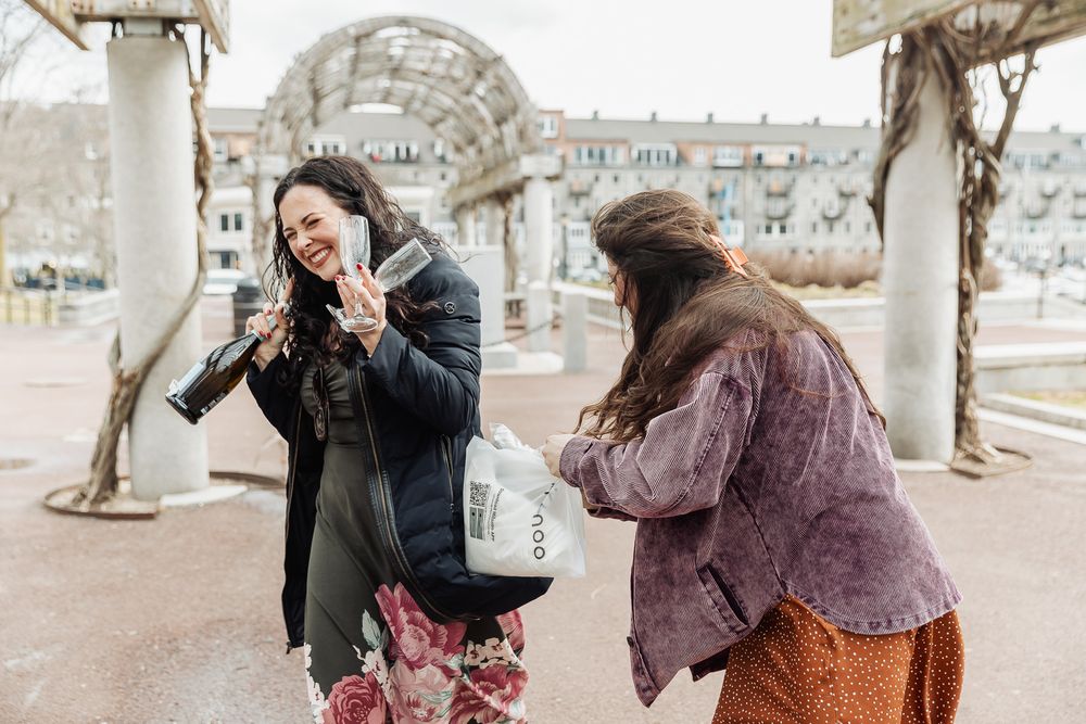 Two women laughing outdoors, one flashing a peace sign while the other looks into a shopping bag.