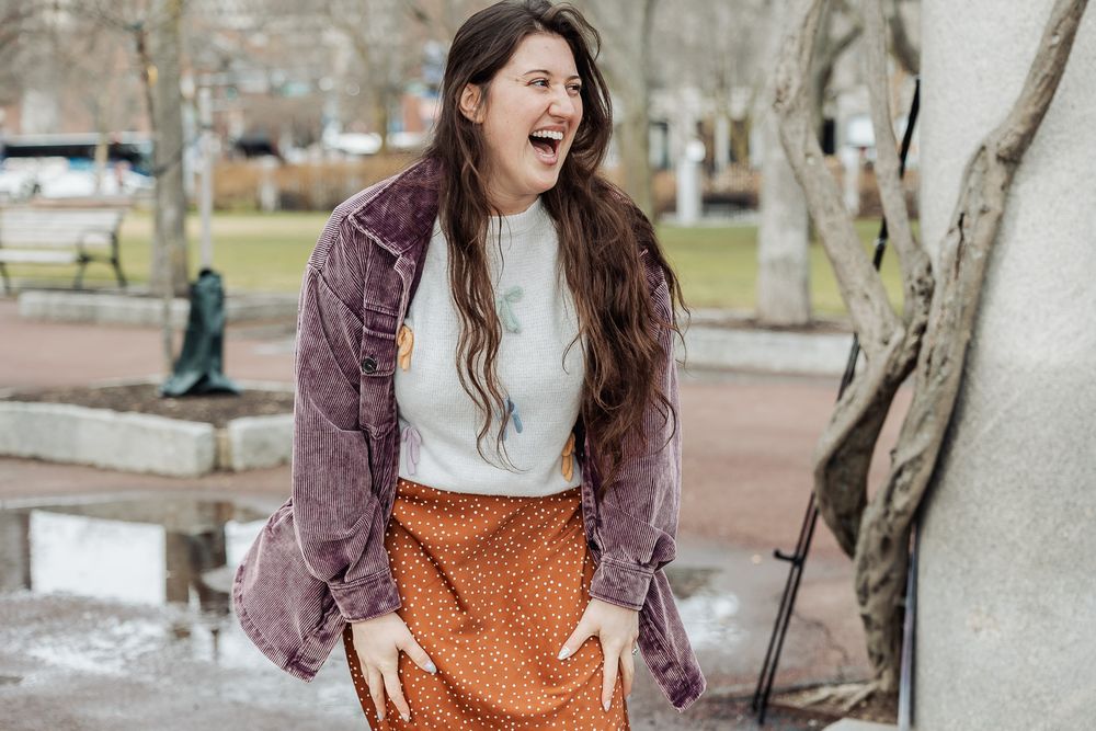 A joyful young woman laughing outdoors in a park, wearing an orange skirt and purple corduroy jacket.