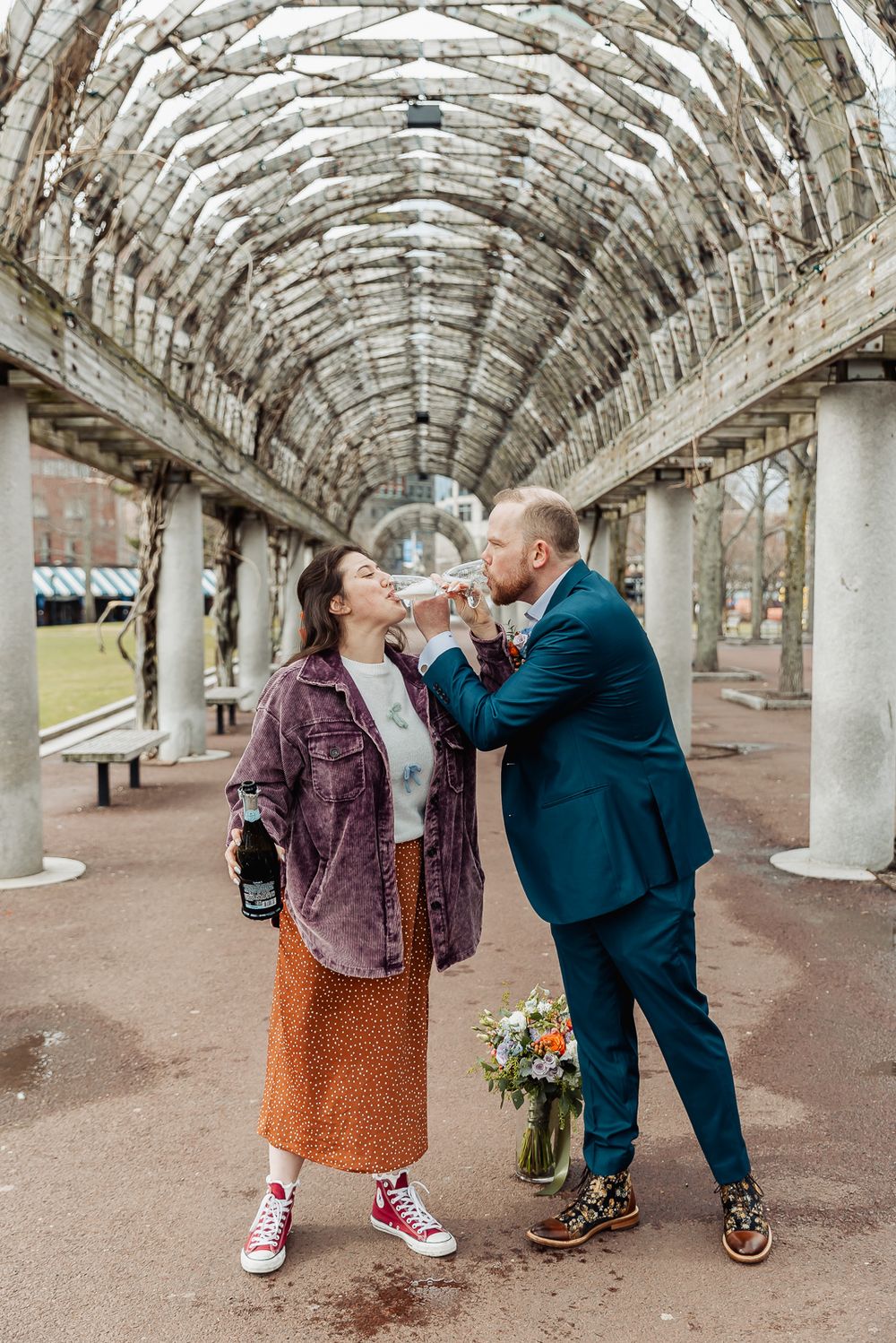 Couple sharing champagne under an arched pergola, man in teal suit holding flowers, woman in denim jacket and orange skirt.